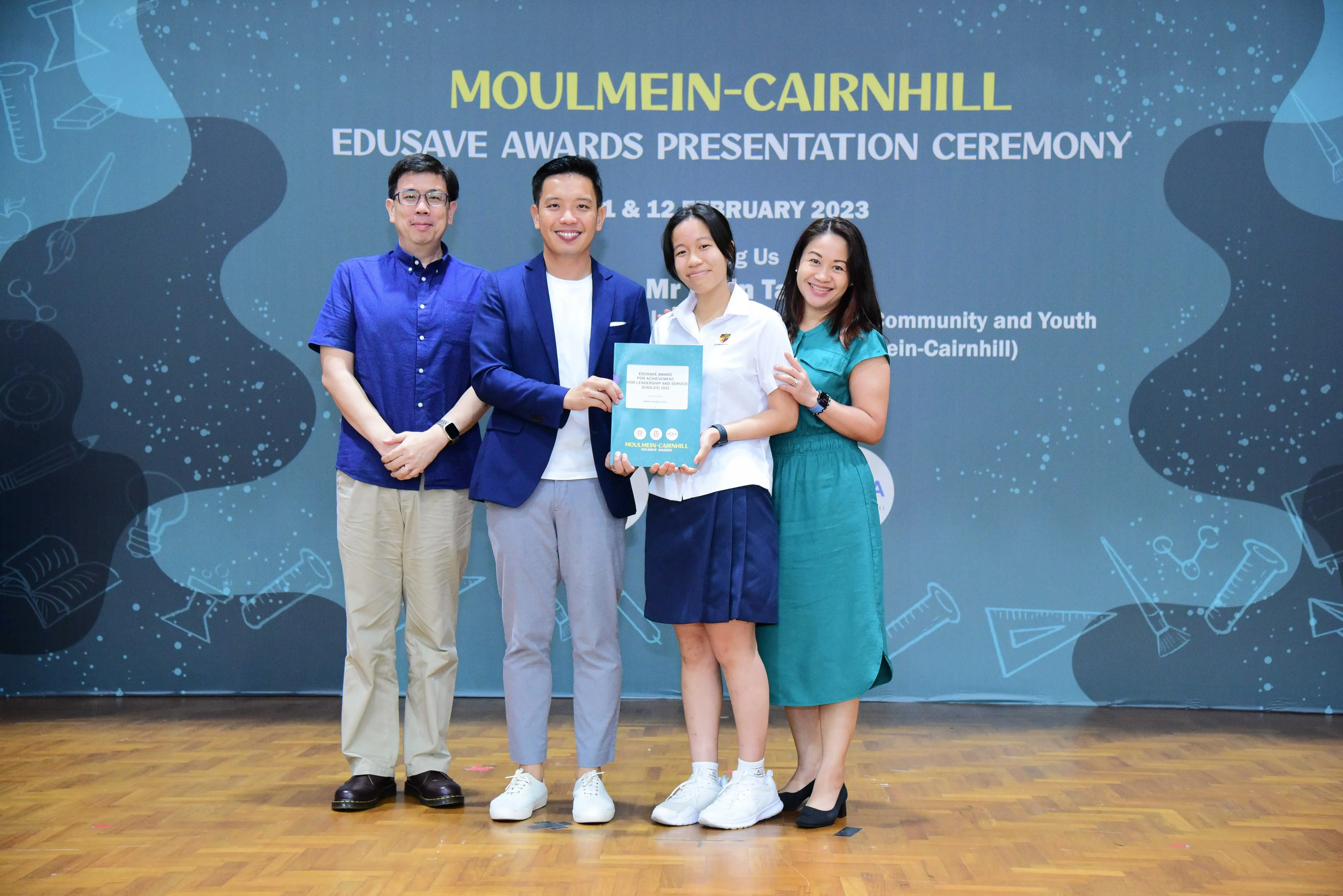 Four people pose at the Moulmein-Cairnhill Edusave Awards Presentation Ceremony, with a student holding a certificate.