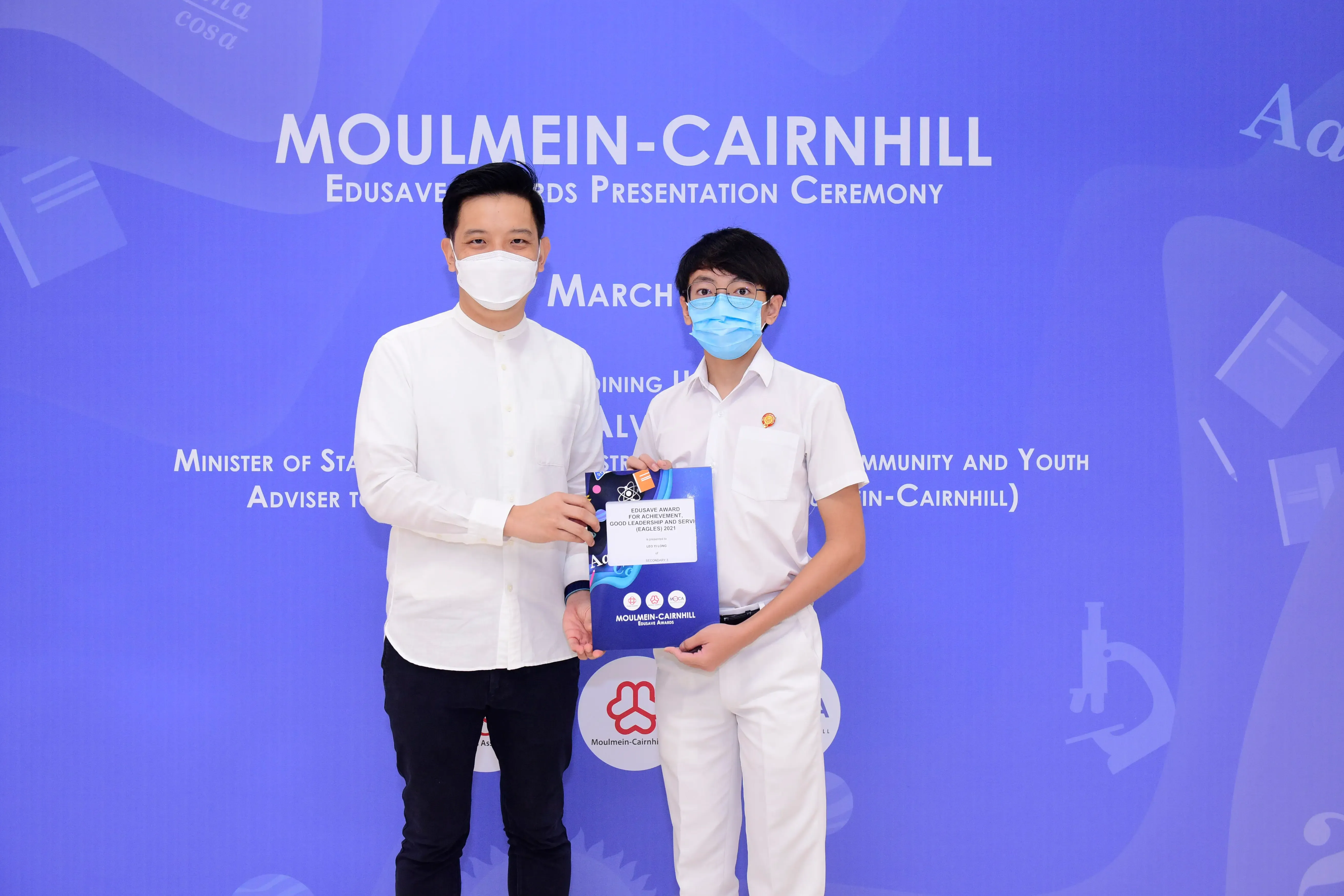 Two masked individuals hold an award certificate at the Moulmein-Cairnhill Edusave Awards Presentation Ceremony.