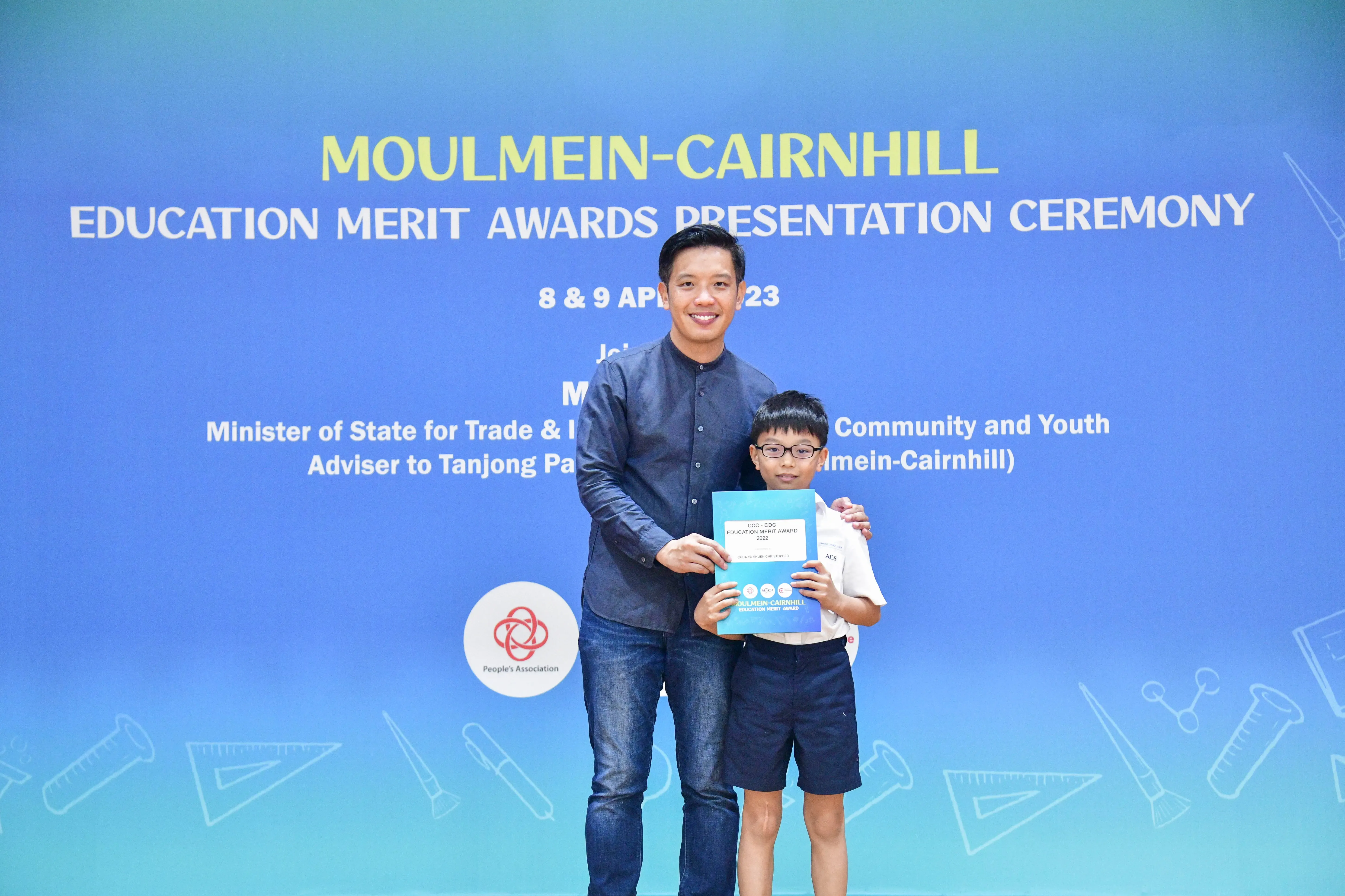 Man and young boy holding a certificate in front of Moulmein-Cairnhill Education Merit Awards Presentation Ceremony banner.