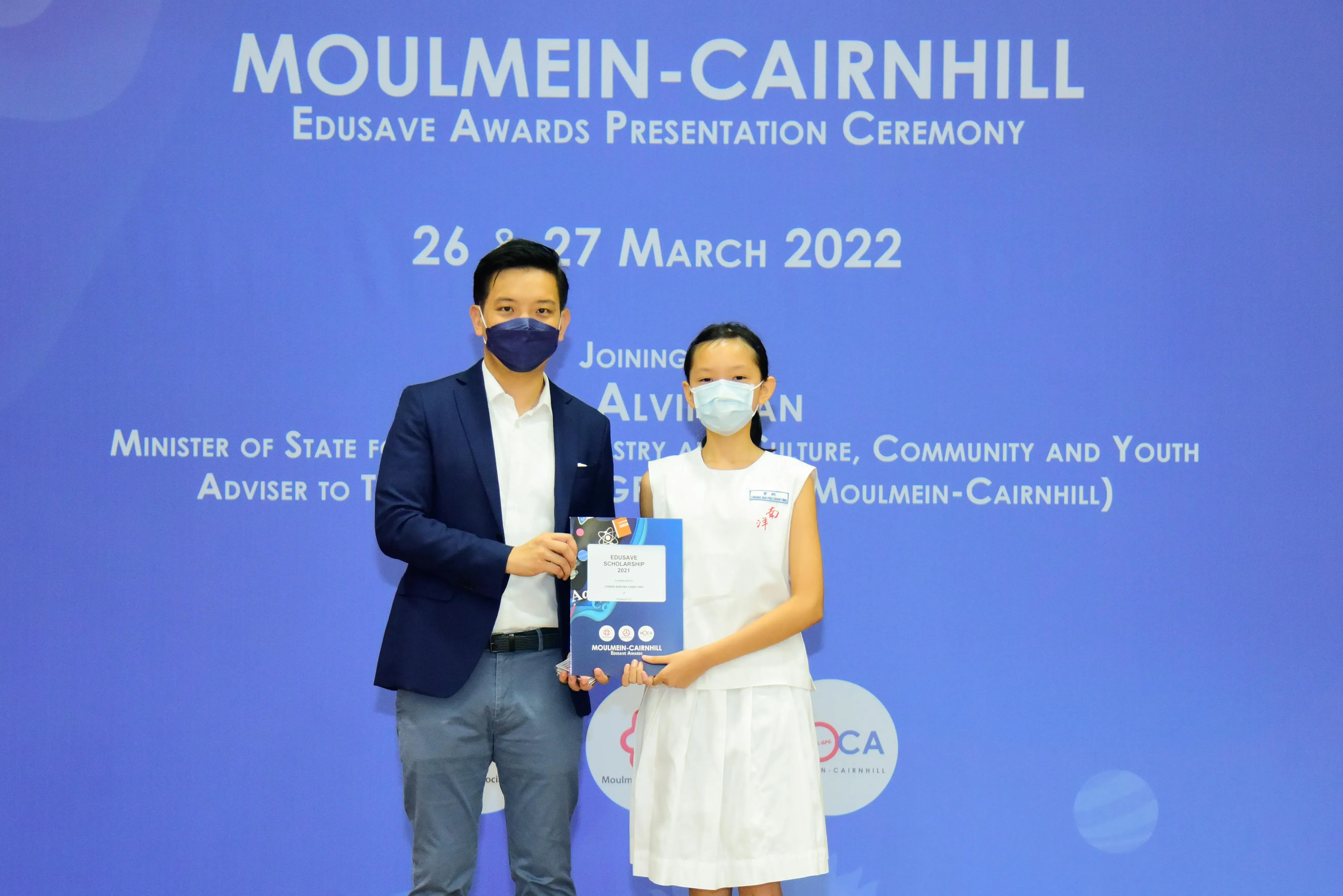 A masked man and young girl hold an Edusave Scholarship at the Moulmein-Cairnhill Awards Ceremony, March 2022.