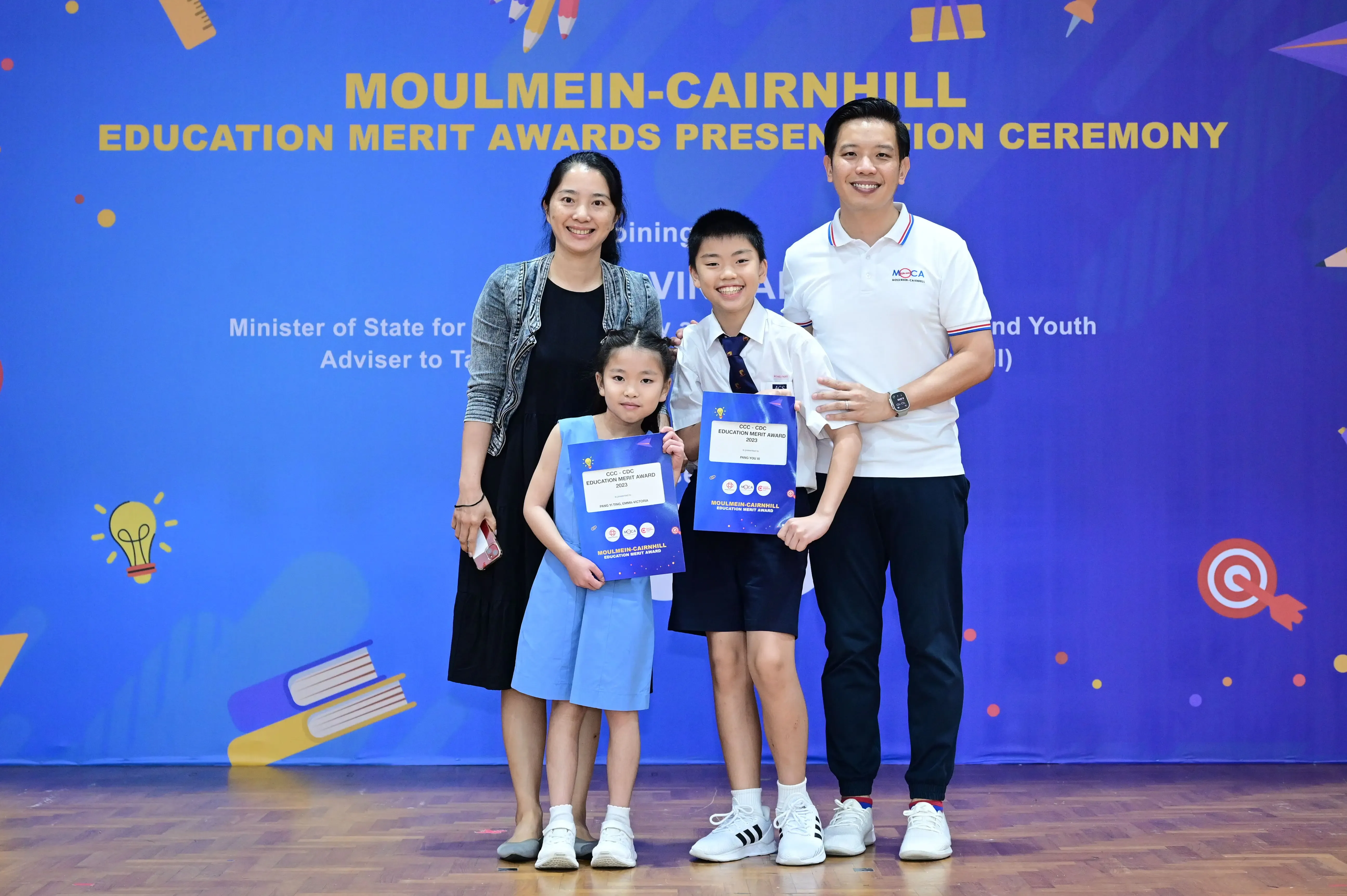 Two adults and two children holding award certificates at the Moulmein-Cairnhill Education Merit Awards ceremony.