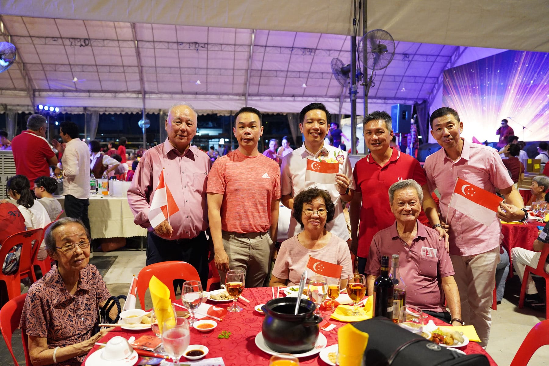 A group of people posing at a festive dinner under a tent, holding Singapore flags, with food on a red table.