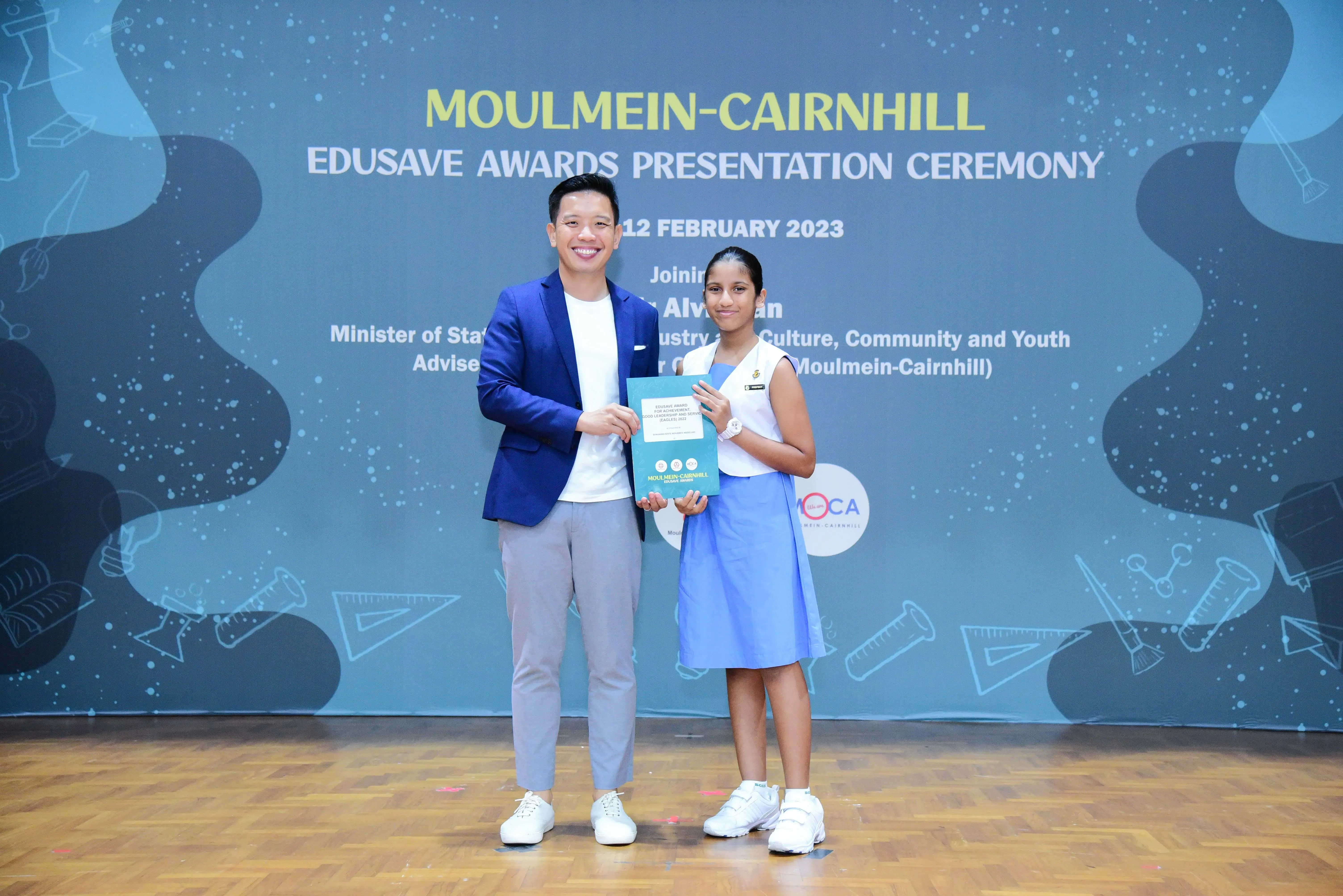 A man in a blue blazer and a young girl in a blue uniform hold a certificate at the Moulmein-Cairnhill Edusave Awards 2023.