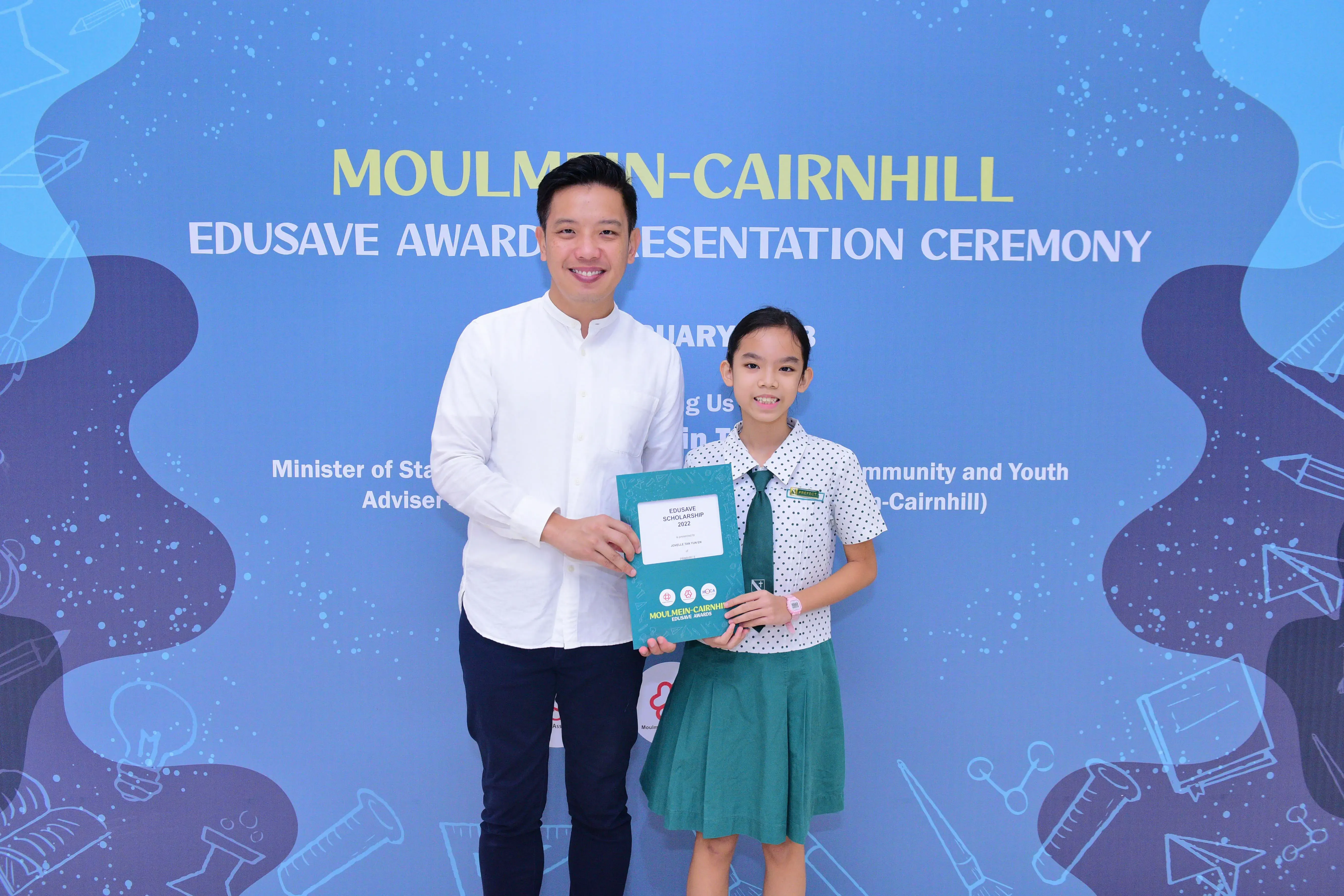A man and a young girl in school uniform hold a certificate at the Moulmein-Cairnhill Edusave Awards Ceremony.