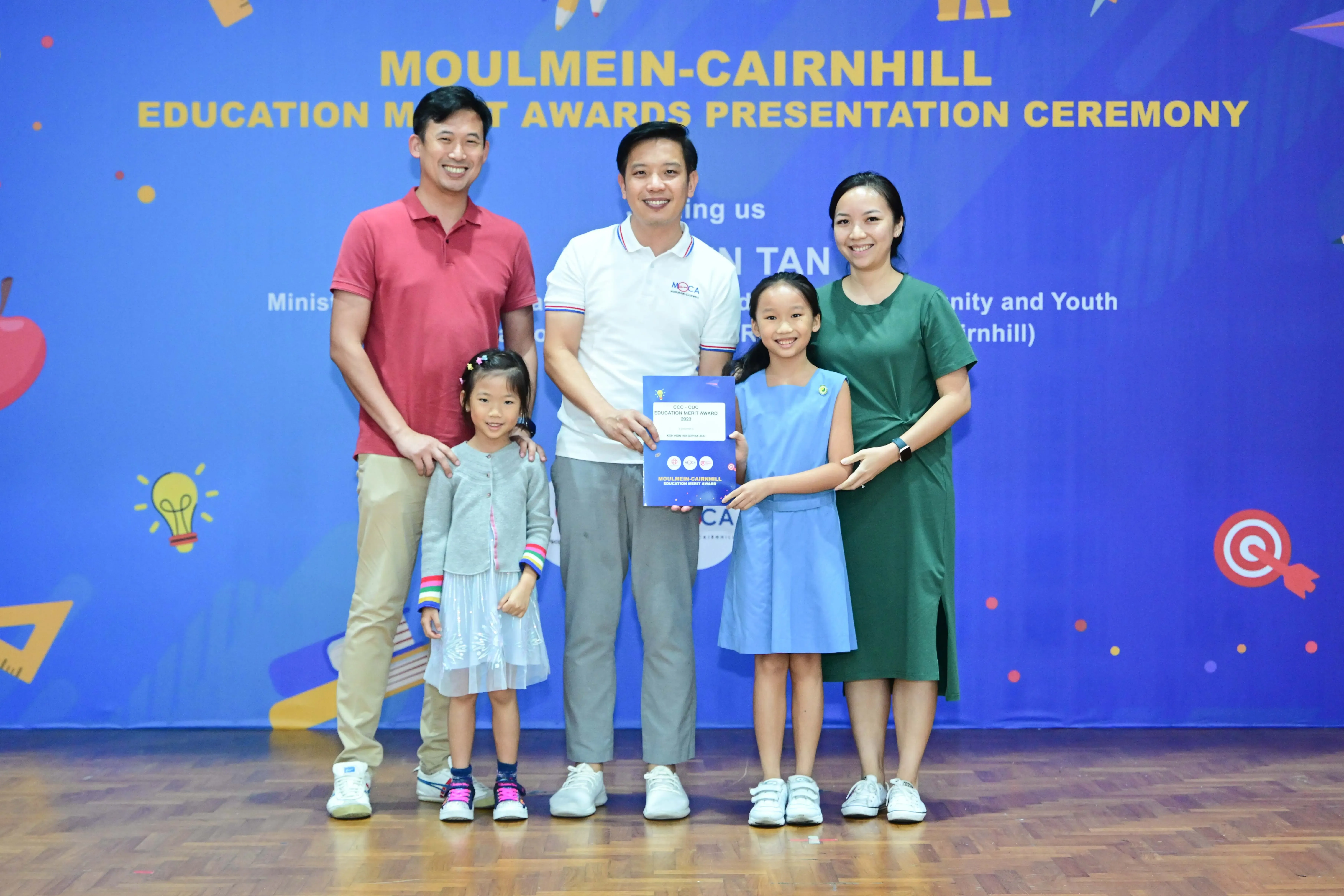 Five people pose at the Moulmein-Cairnhill Education Merit Awards Presentation Ceremony, holding a blue award certificate.