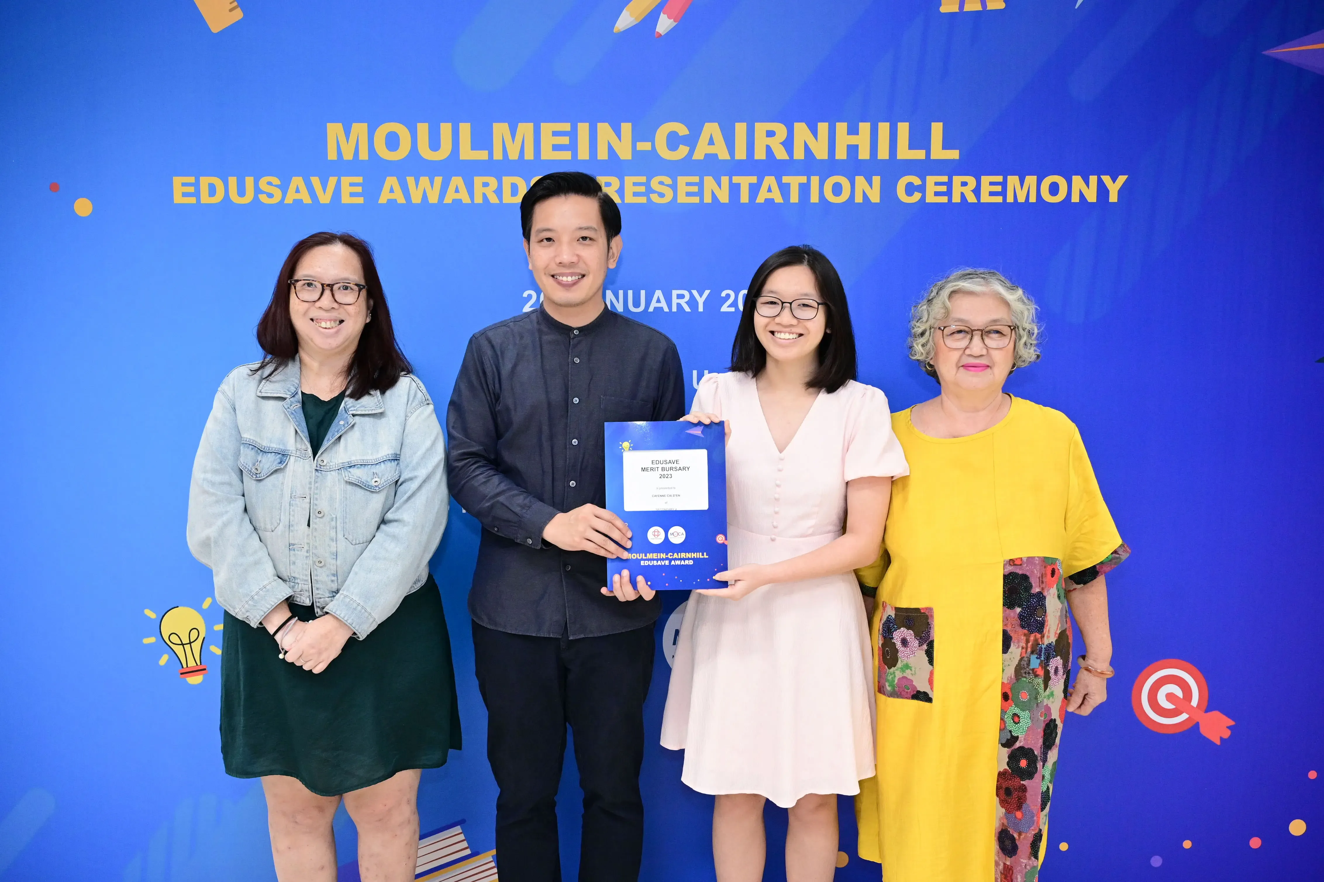 Four people smile in front of a blue Moulmein-Cairnhill Edusave Awards Presentation Ceremony banner, holding a certificate.