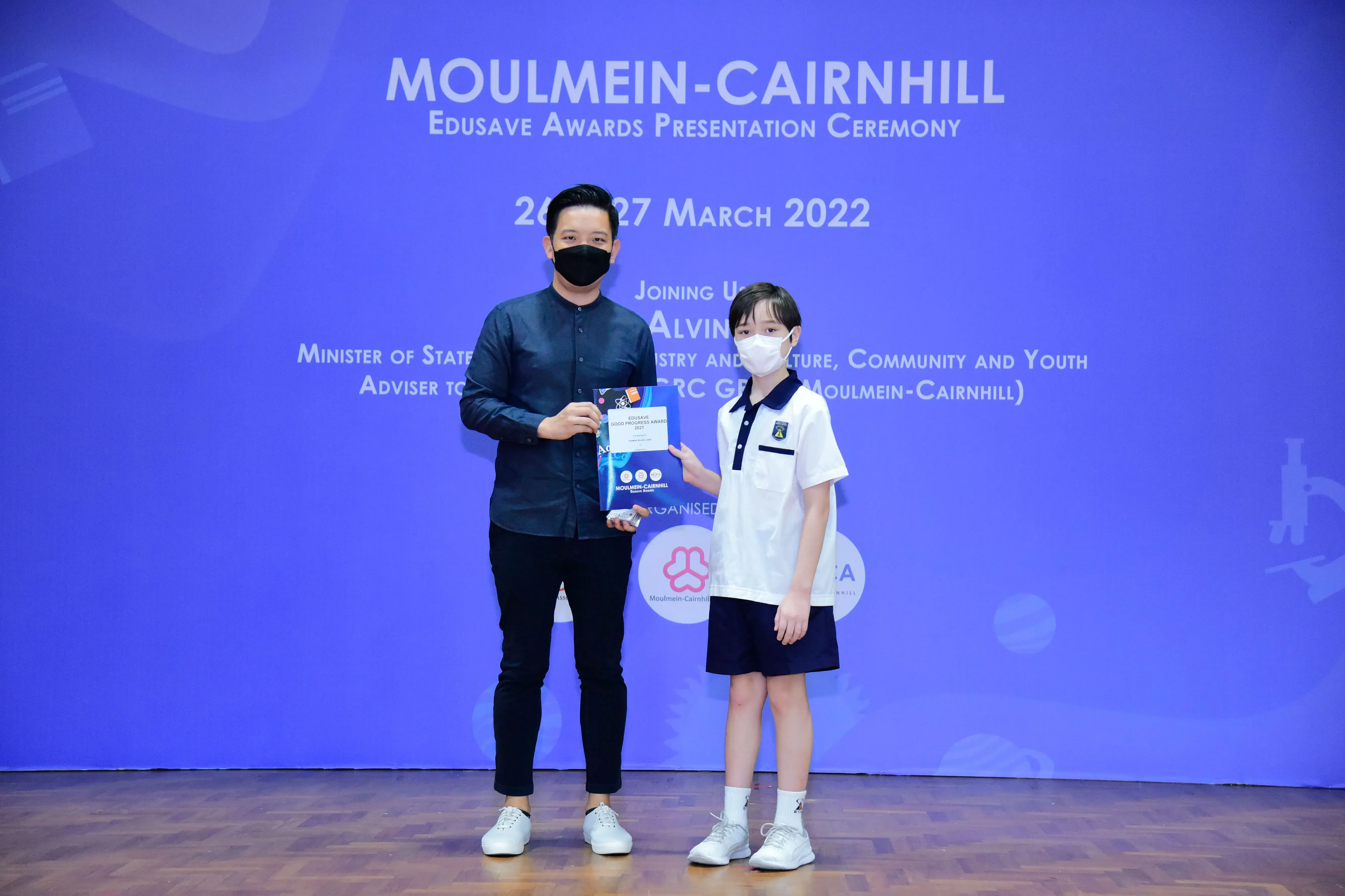 An adult and a young student in uniform hold a certificate at the Moulmein-Cairnhill Edusave Awards Ceremony, March 2022.