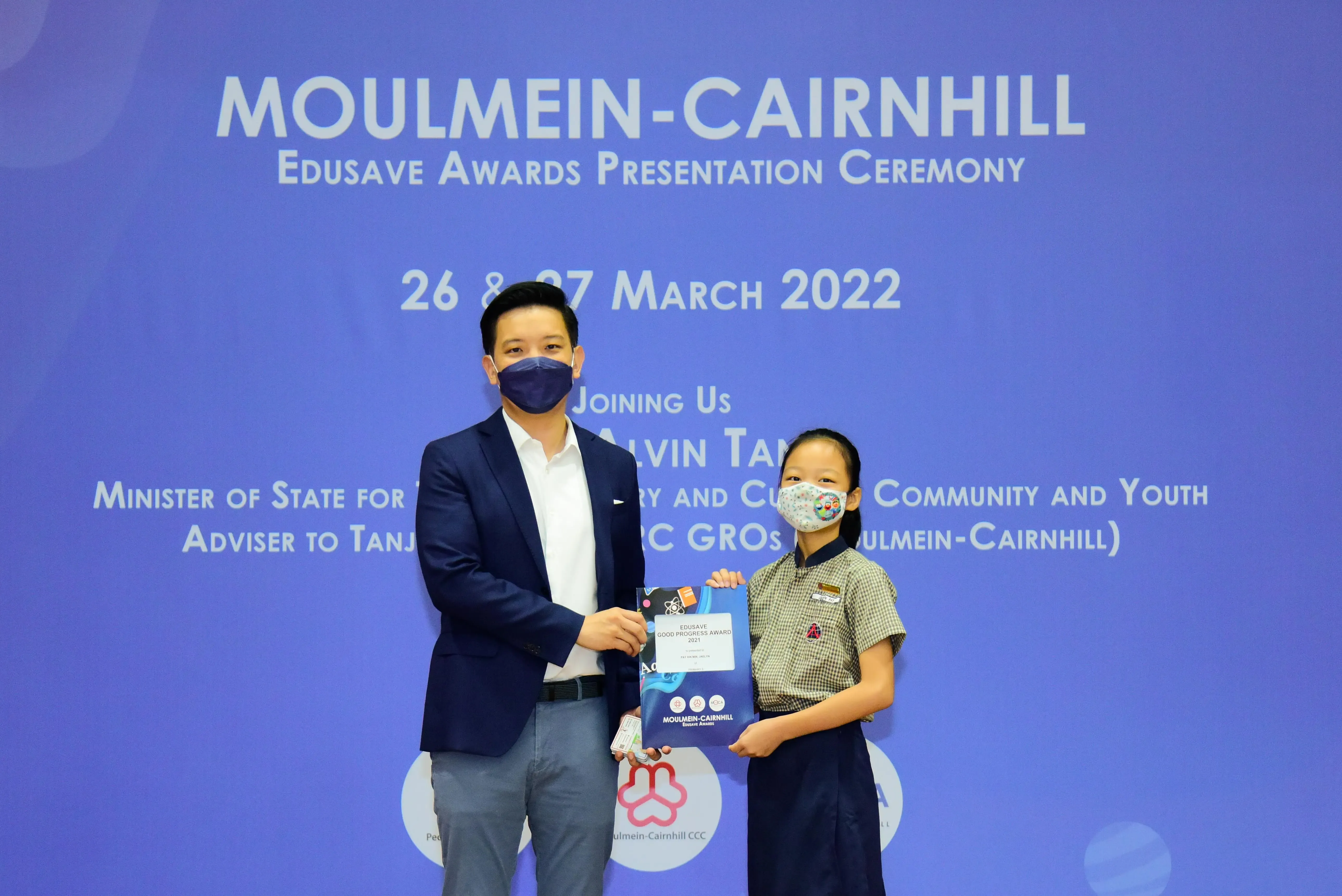 Man in a suit and a young girl in school uniform hold an award certificate in front of a blue ceremony backdrop.