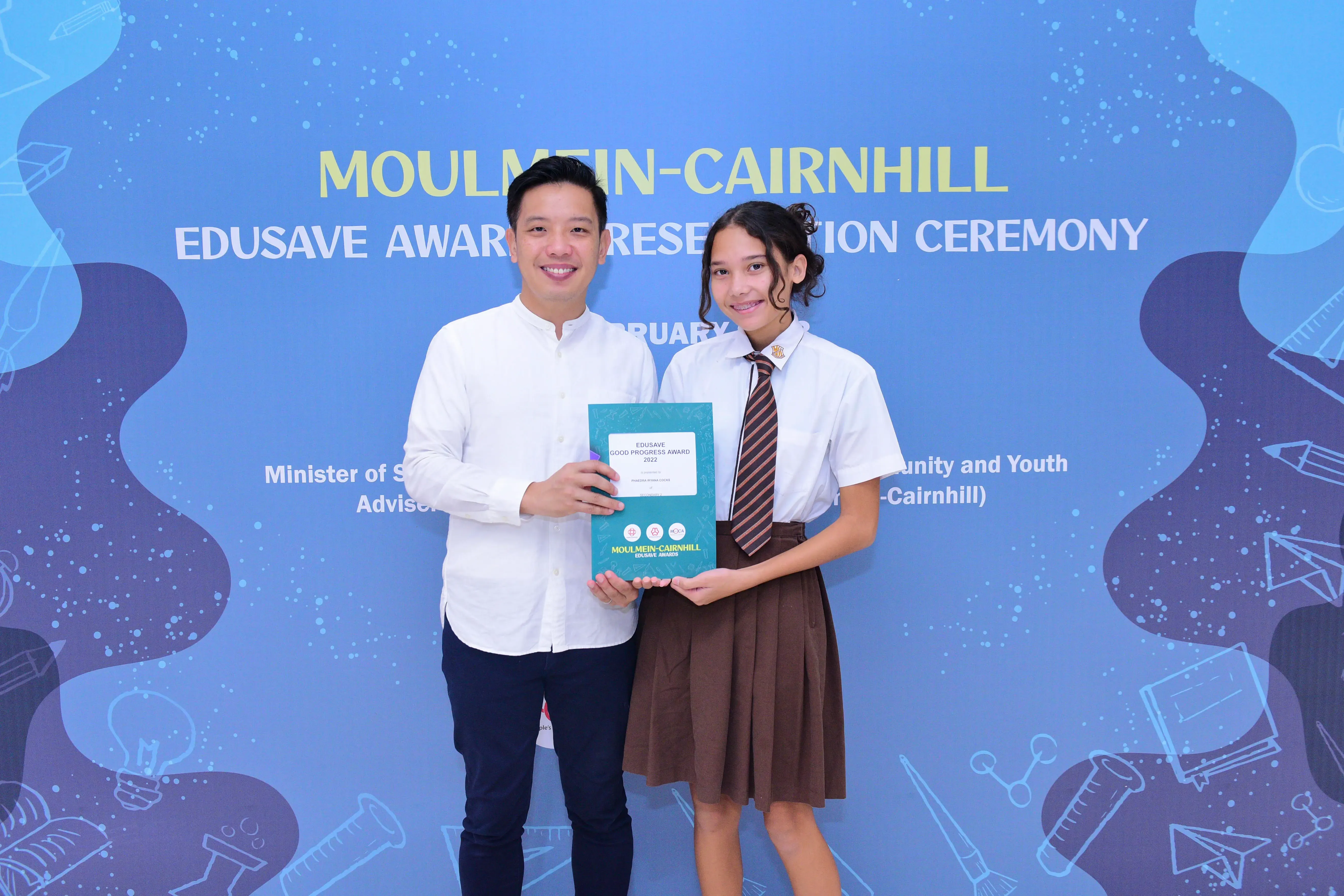 A man and a uniformed student hold an Edusave award certificate at the Moulmein-Cairnhill Edusave Award Presentation Ceremony.