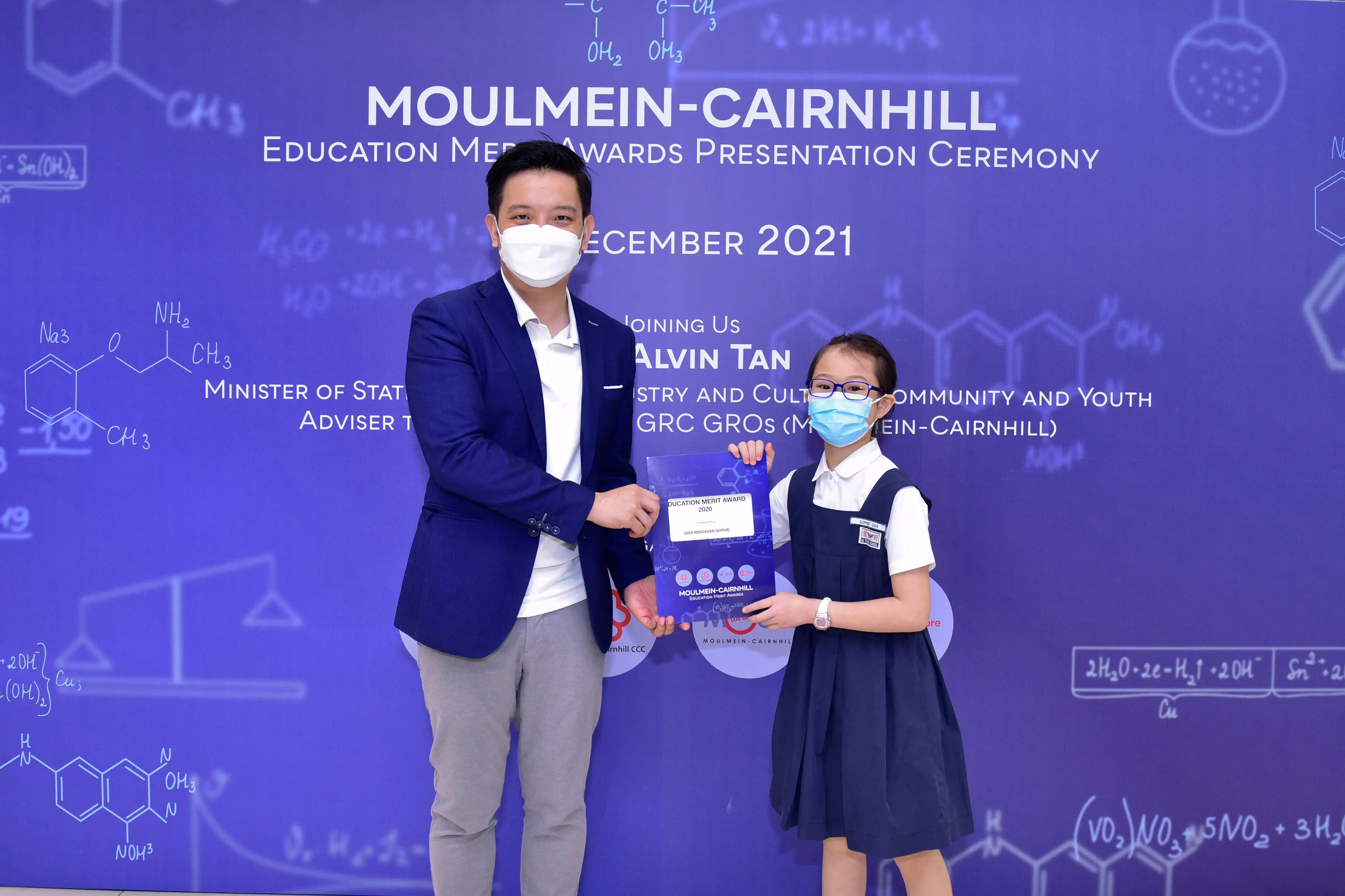 A masked man presents an award certificate to a masked young girl at the Moulmein-Cairnhill Education Merit Awards 2021.