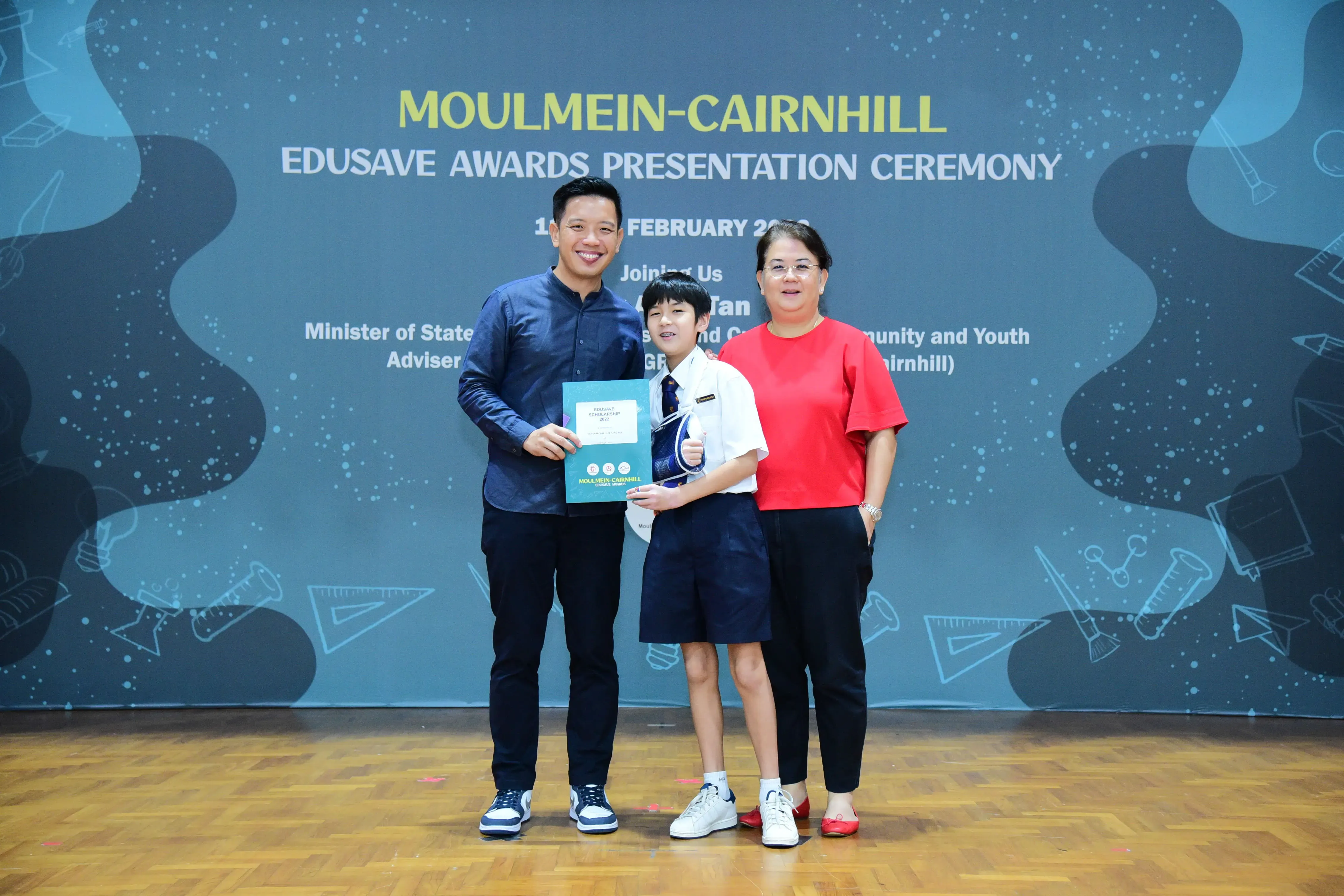 A young student in school uniform holds a certificate between two adults at the Moulmein-Cairnhill Edusave Awards Ceremony.