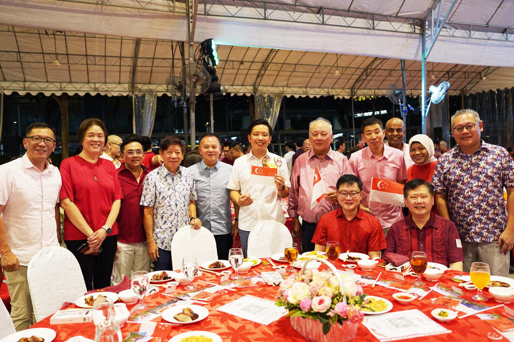 A group of people posing and smiling at a dinner event with Singapore flags and food on a red-clothed table.