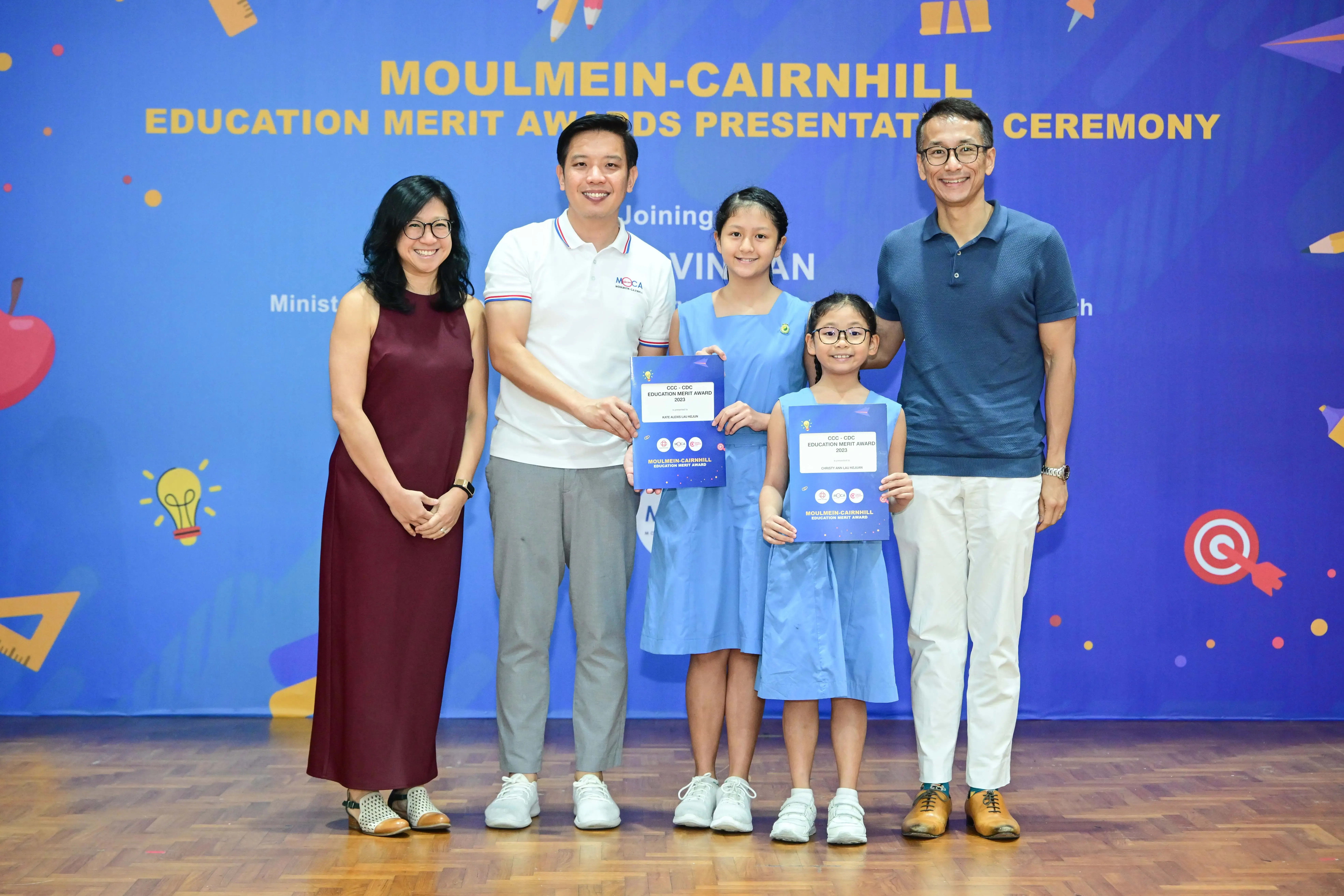 Five people pose at the Moulmein-Cairnhill Education Merit Awards, with two girls holding blue award certificates.