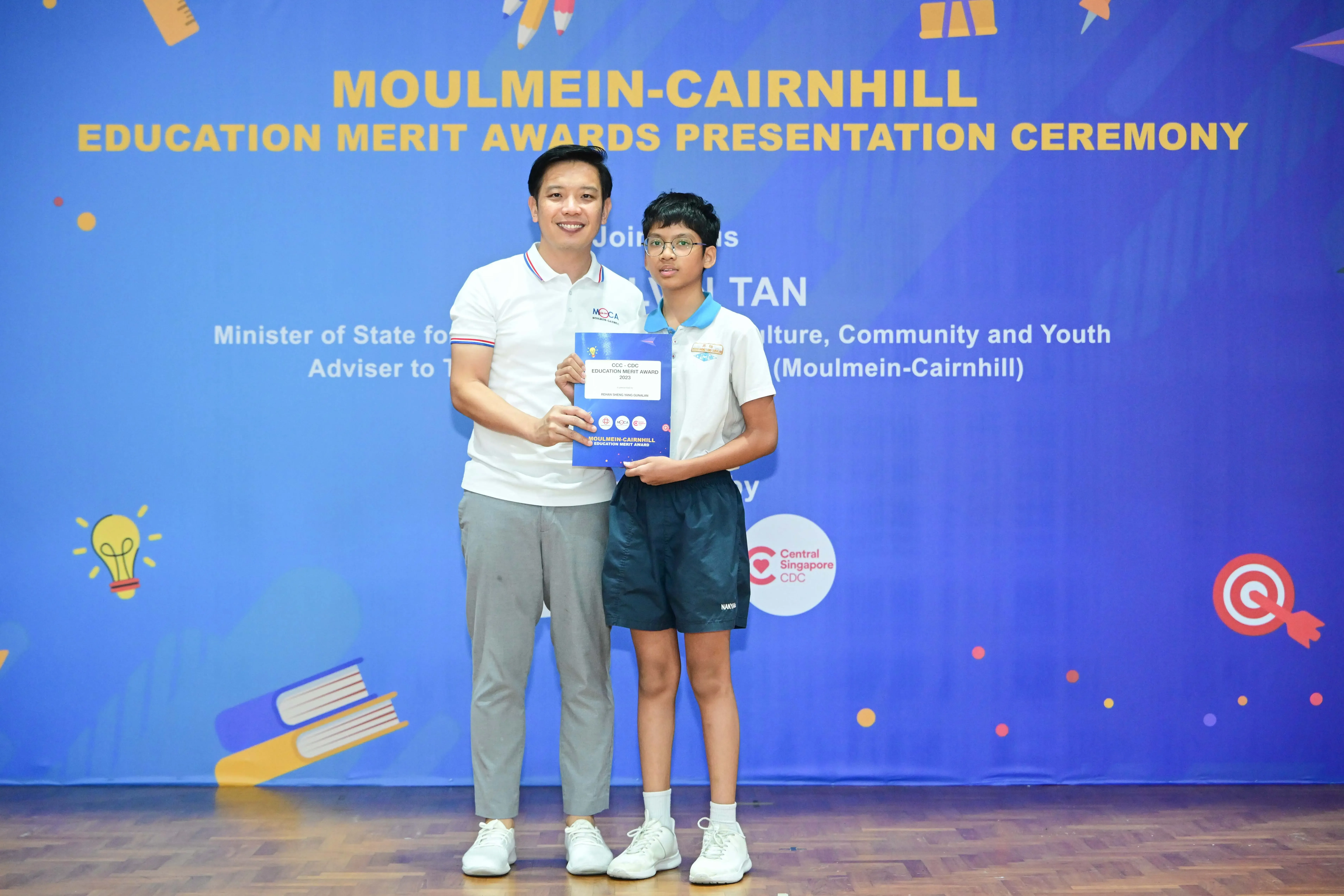 An adult and a young student hold an award certificate at the Moulmein-Cairnhill Education Merit Awards ceremony.