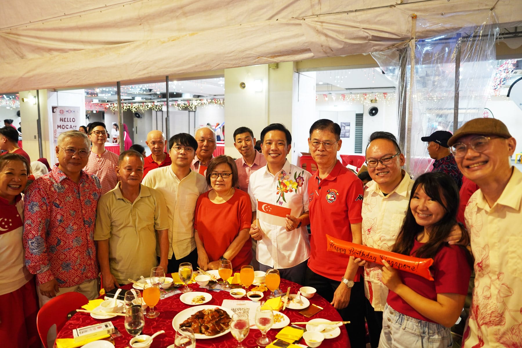 Group of people in red outfits posing at a Singapore National Day dinner, holding a small flag and a Happy Birthday Singapore balloon.