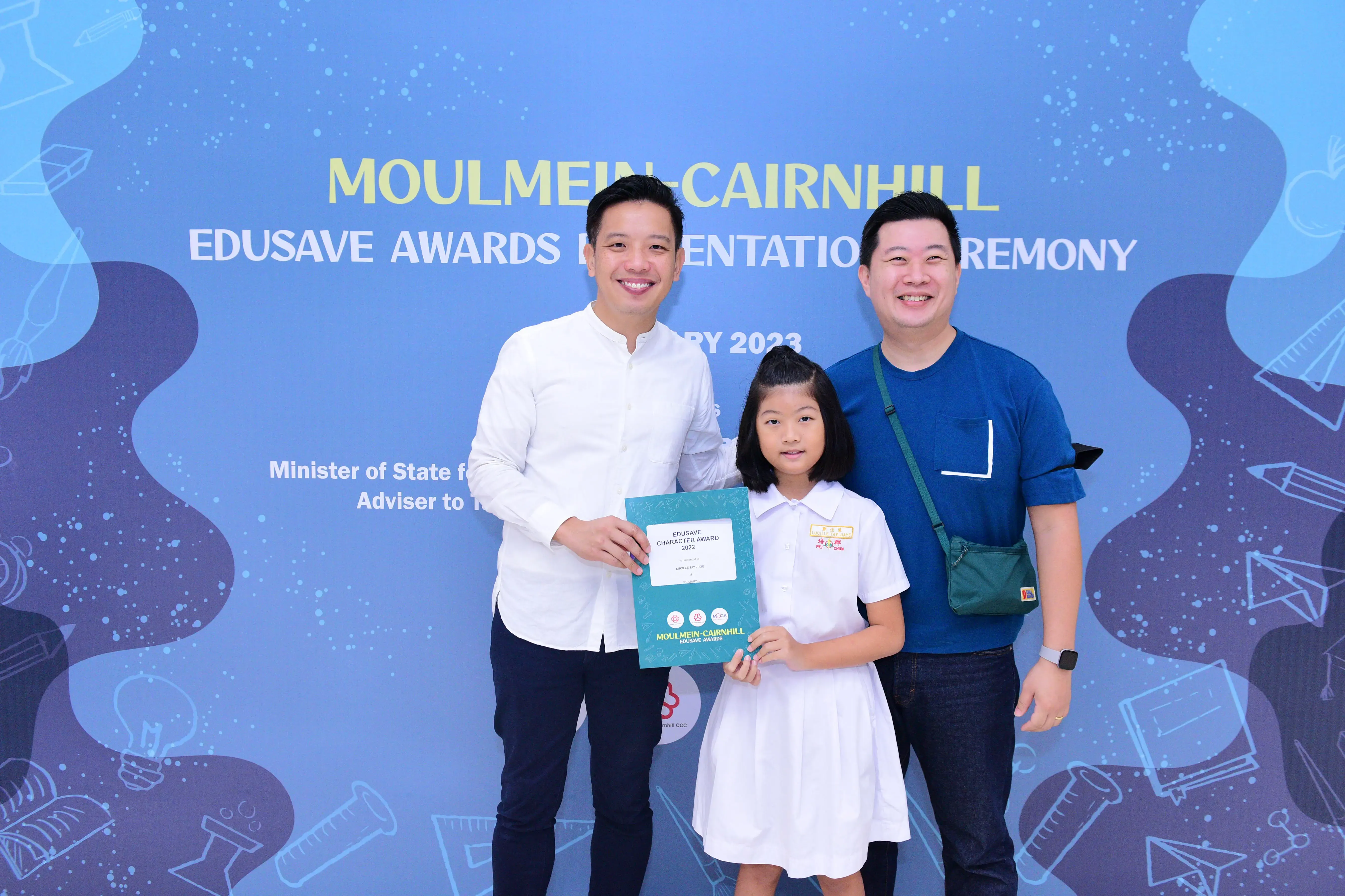 Two men and a young girl pose holding an Edusave Character Award certificate at the Moulmein-Cairnhill ceremony 2023.