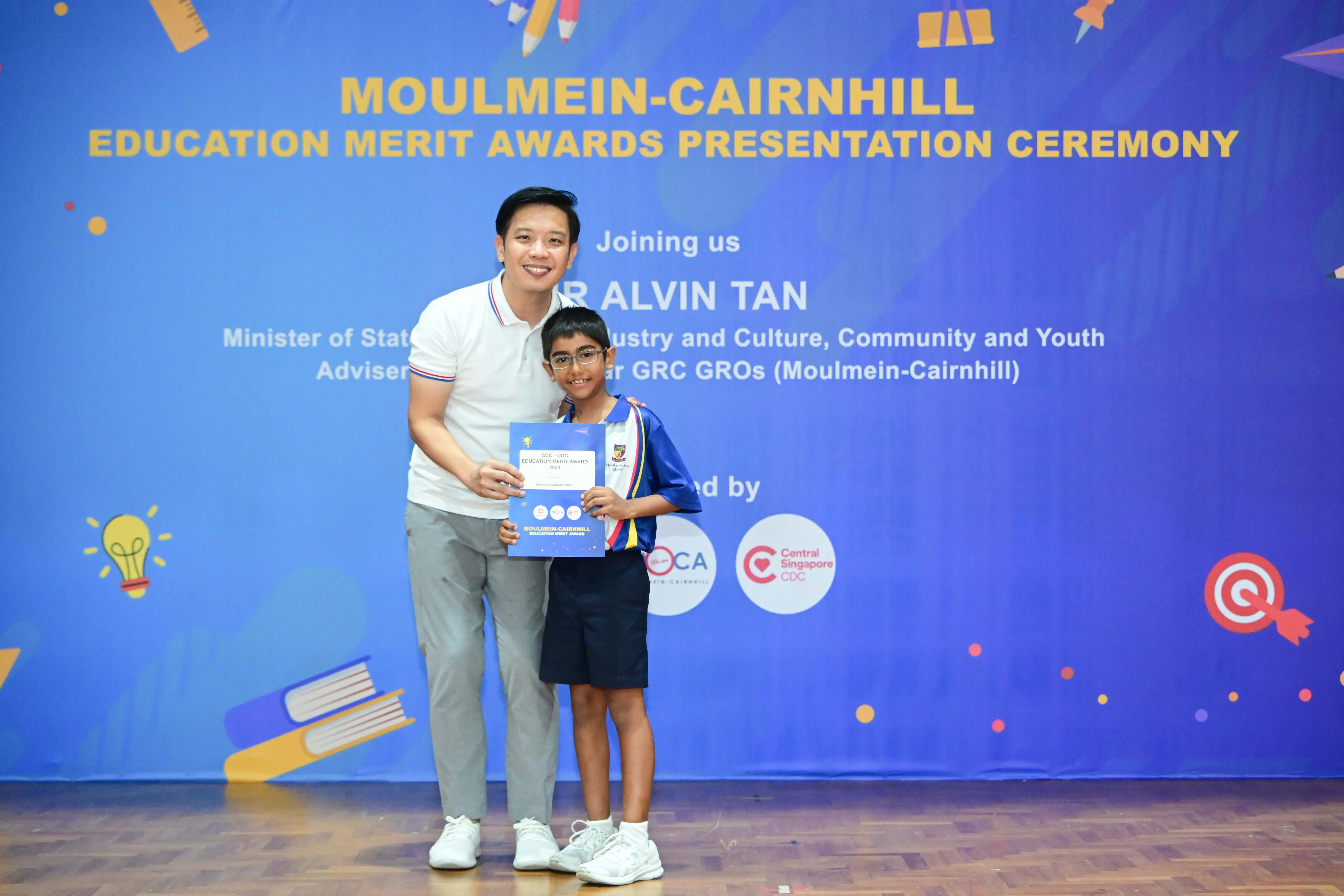 An adult and a young student hold a certificate in front of a Moulmein-Cairnhill Education Merit Awards banner.
