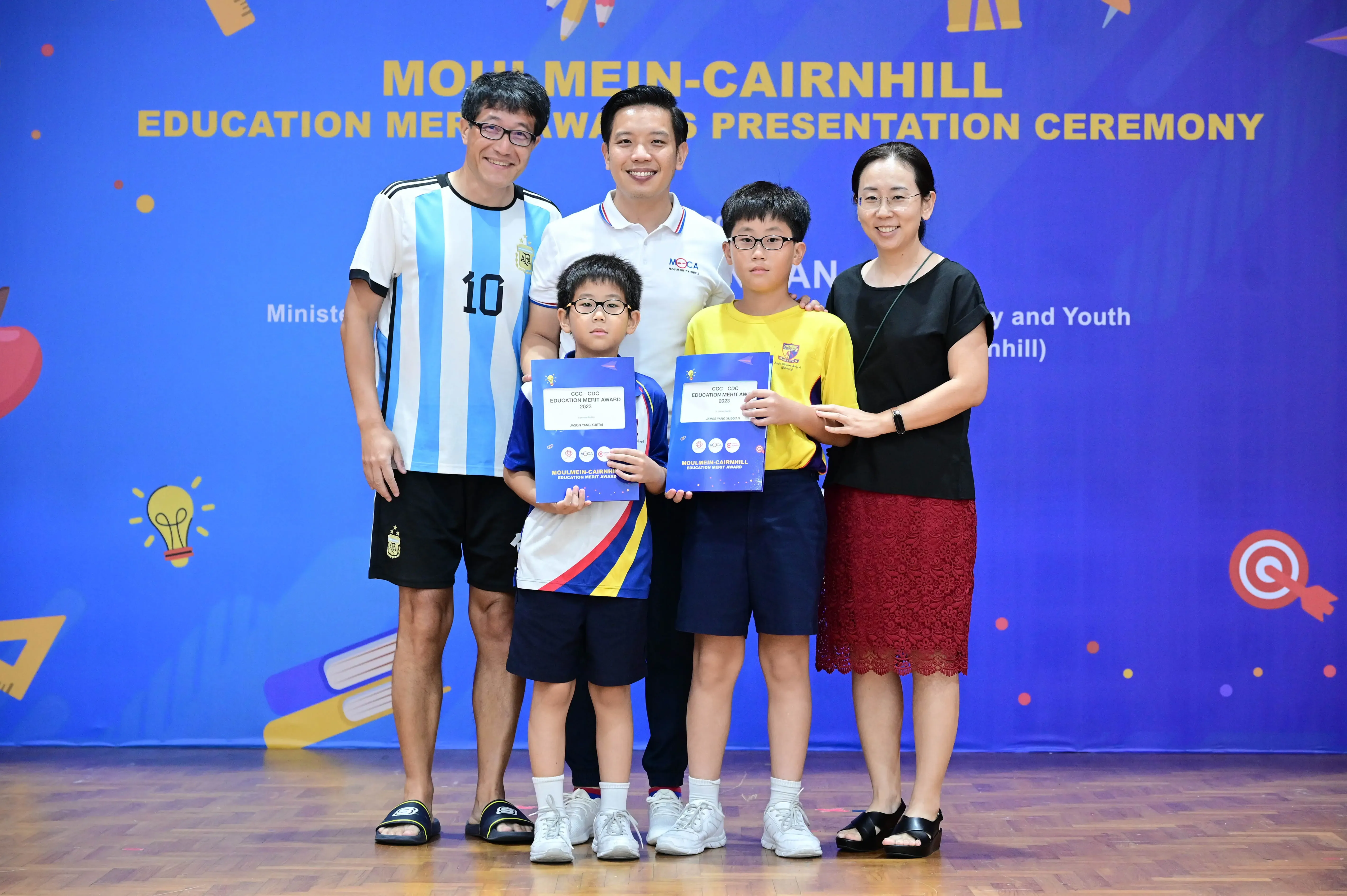 Five people pose before a Moulmein-Cairnhill Education Merit Awards banner, two boys holding blue award certificates.