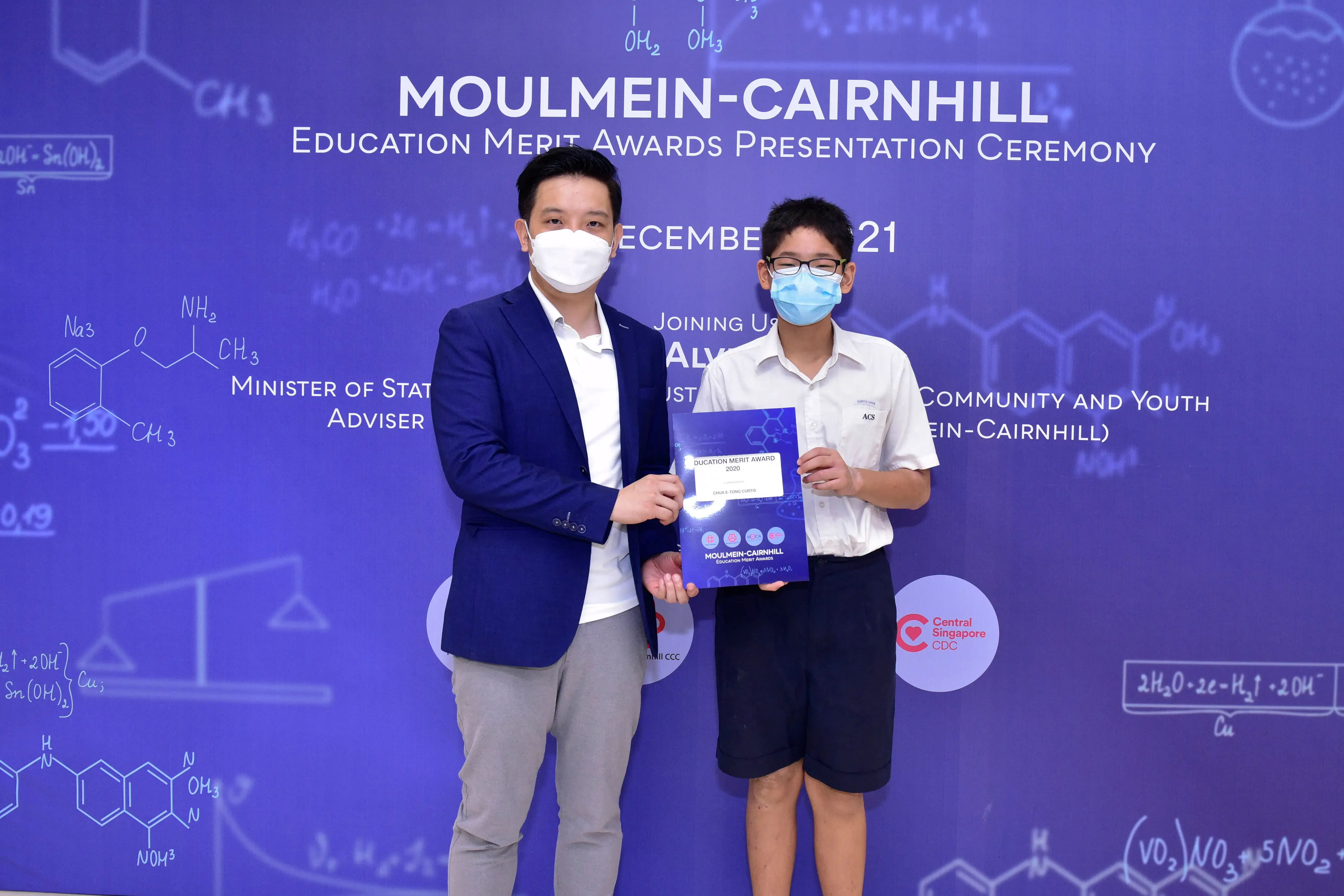 Two masked individuals hold an award certificate at the Moulmein-Cairnhill Education Merit Awards Presentation Ceremony.
