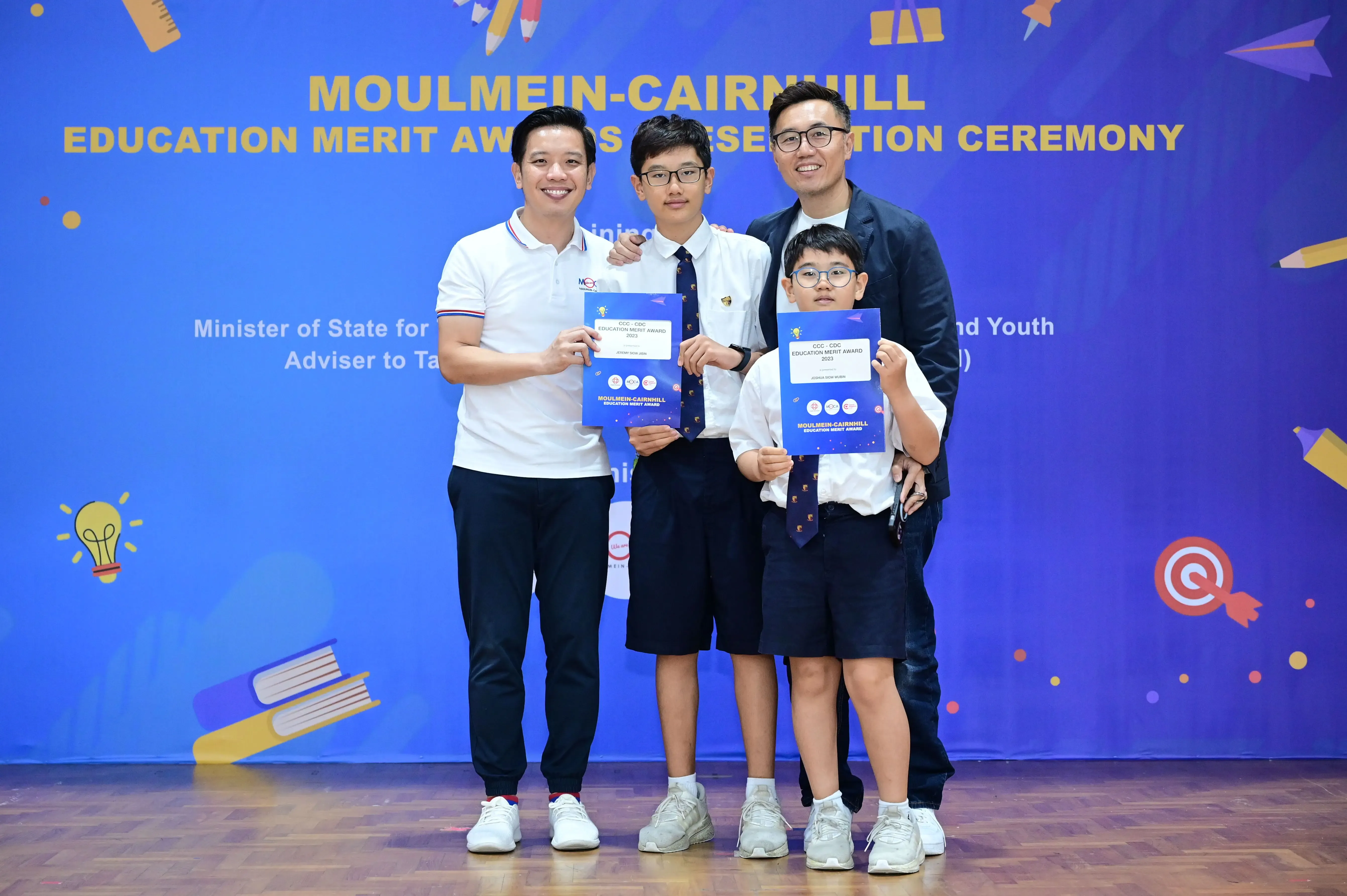 Two adults and two students holding award certificates at the Moulmein-Cairnhill Education Merit Awards Ceremony.