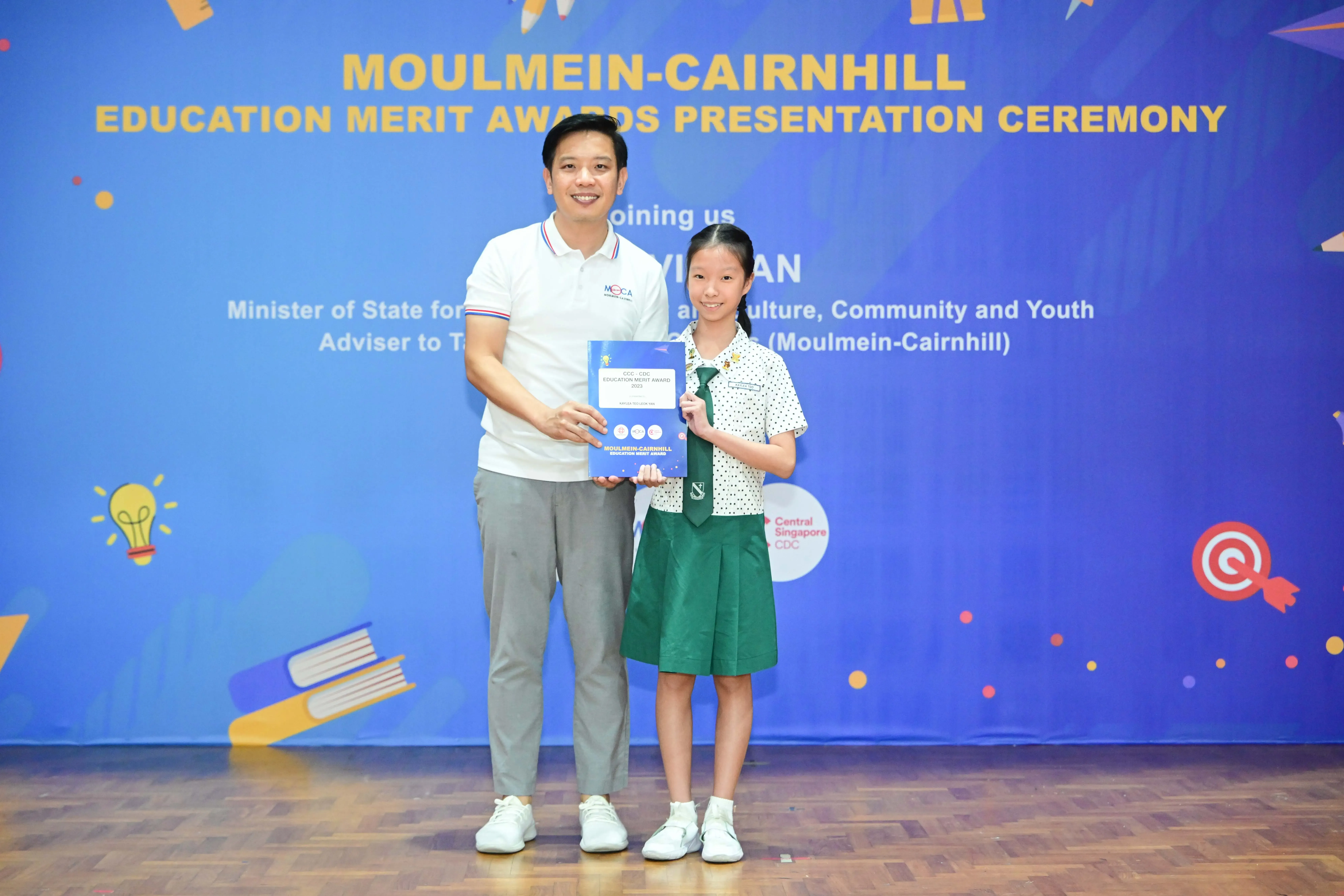 A man and a young girl in school uniform hold an award certificate at the Moulmein-Cairnhill Education Merit Awards ceremony.