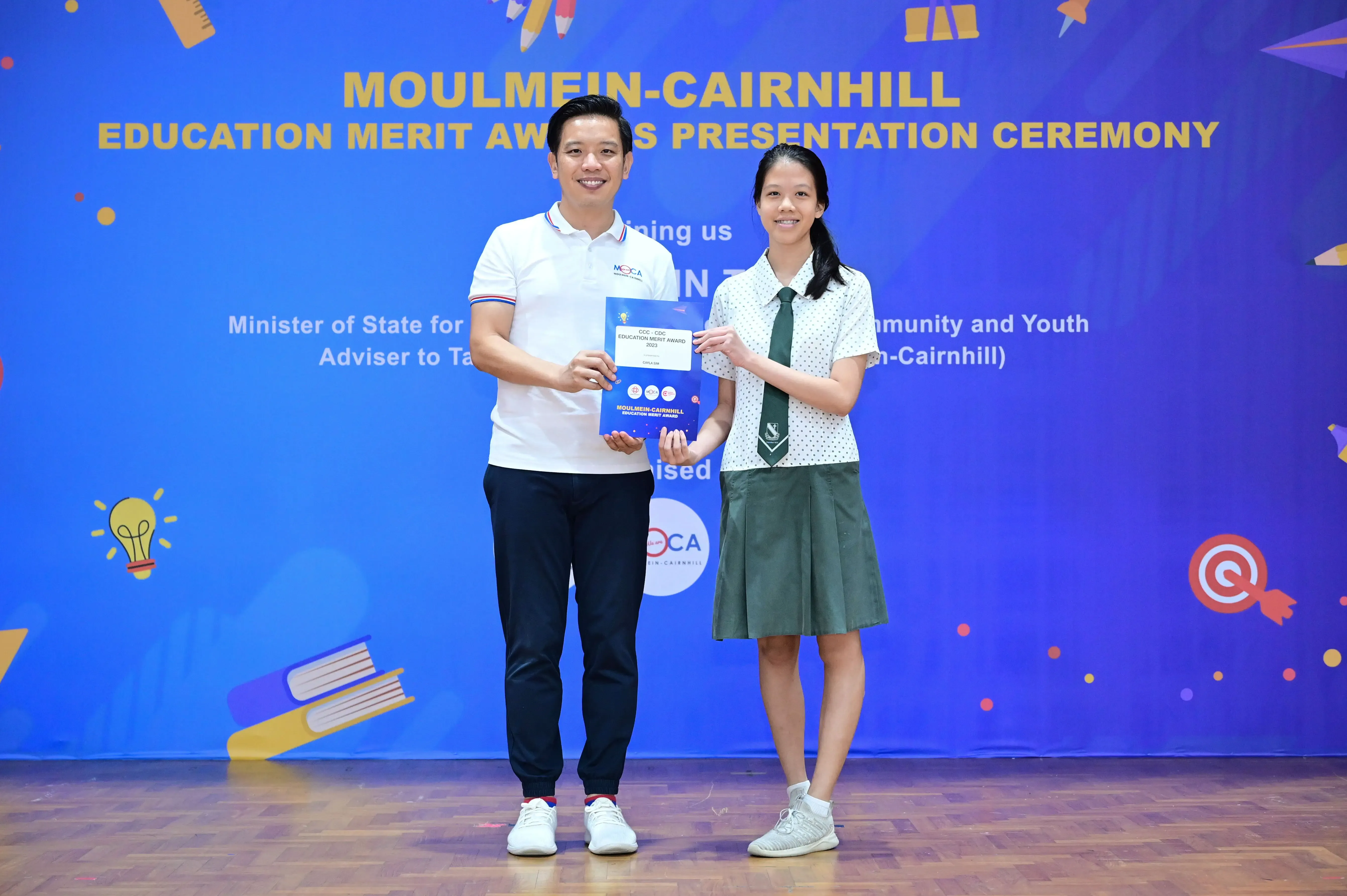 A man and a female student hold an award certificate at the Moulmein-Cairnhill Education Merit Awards Presentation Ceremony.