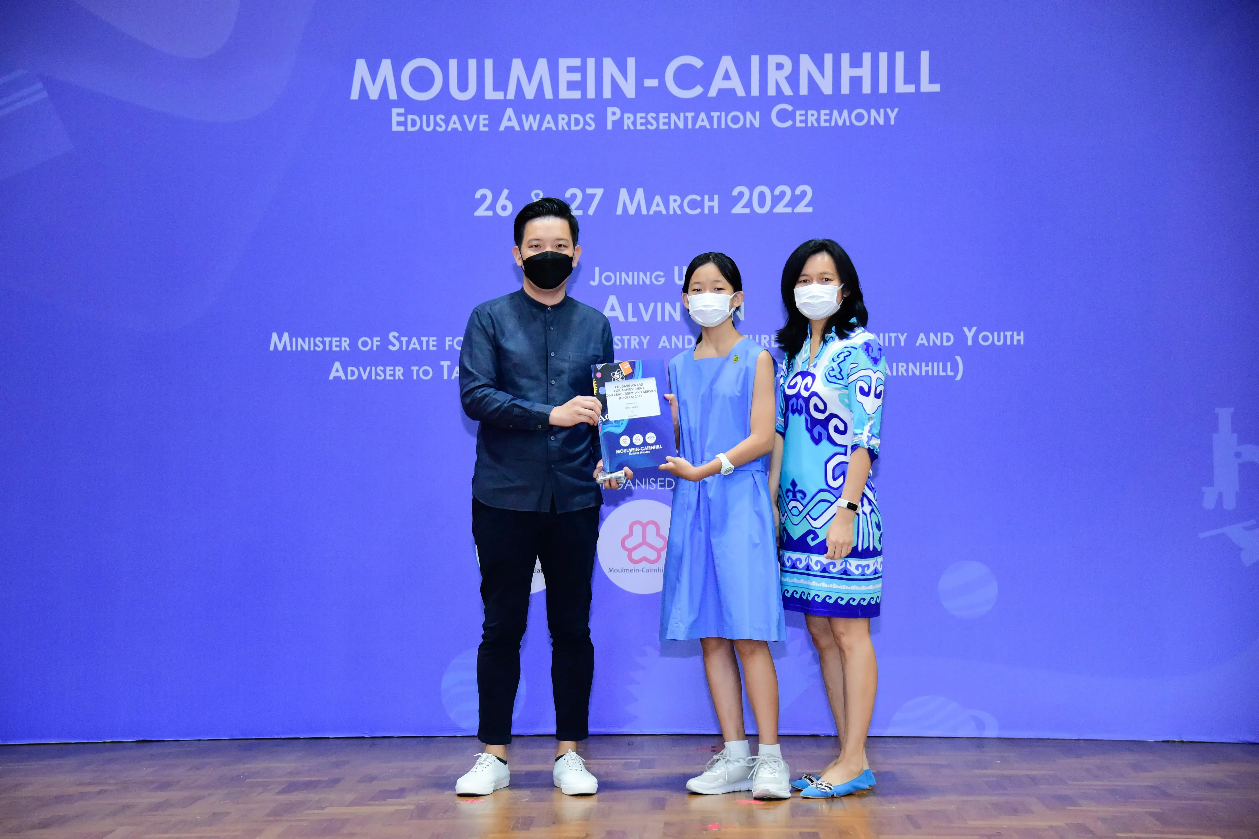 Three masked people pose on stage at the Moulmein-Cairnhill Edusave Awards Presentation Ceremony, March 2022.