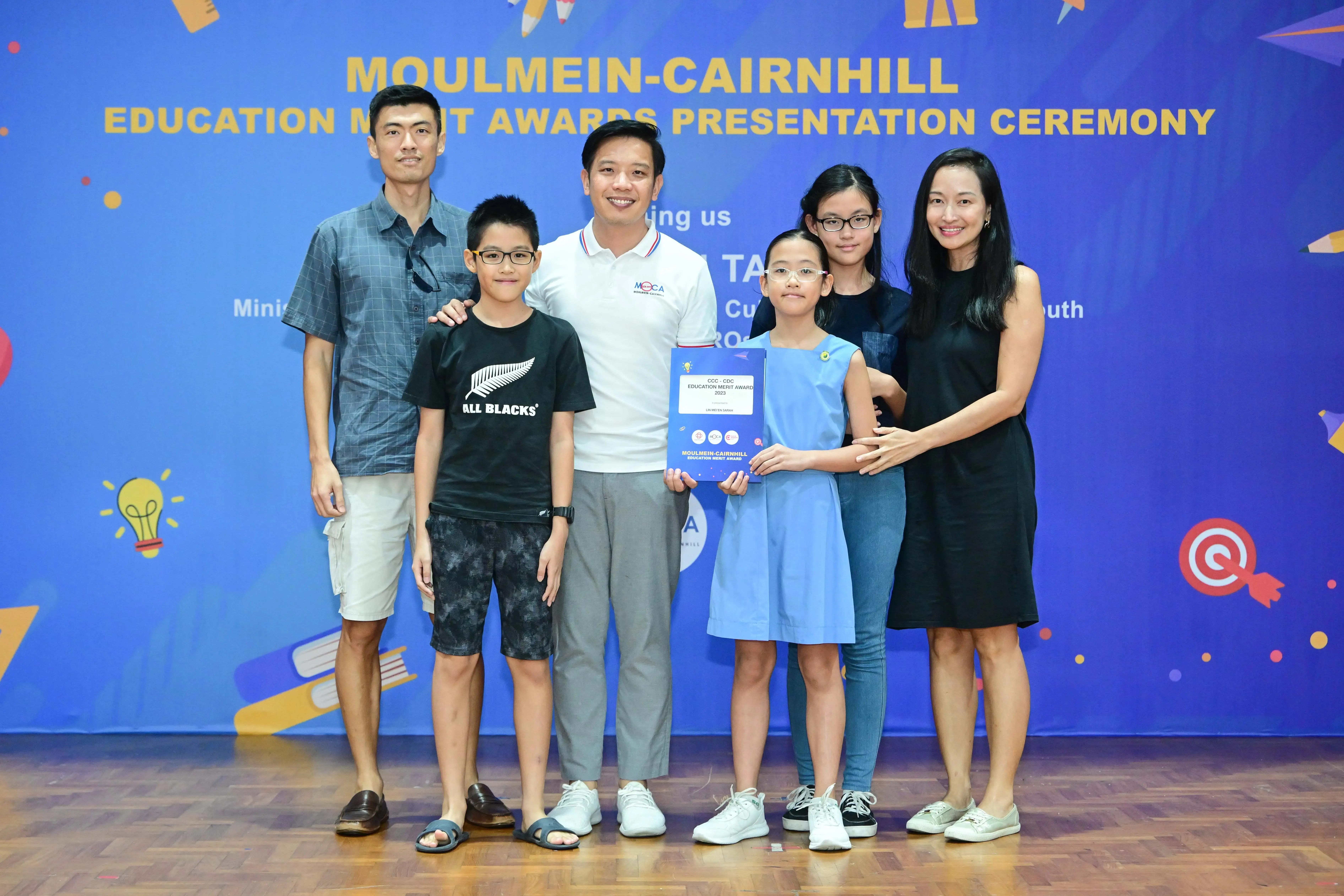 Six people pose in front of a blue Moulmein-Cairnhill Education Merit Awards Presentation Ceremony backdrop.