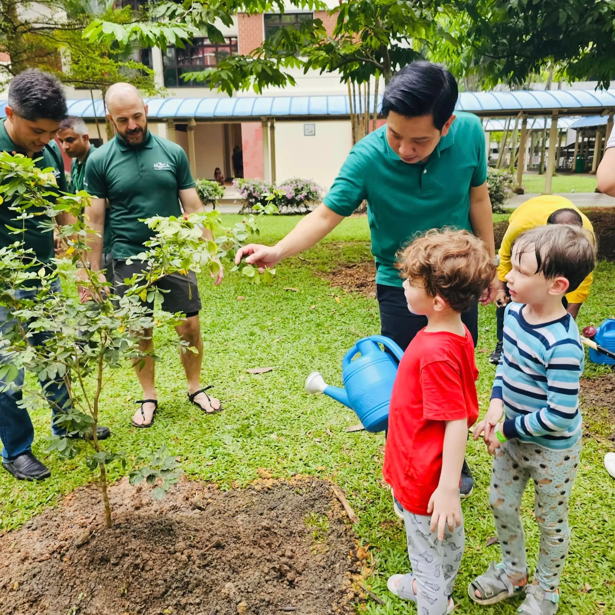 Adults and young children gather outdoors around a freshly planted small tree, with one child holding a blue watering can.