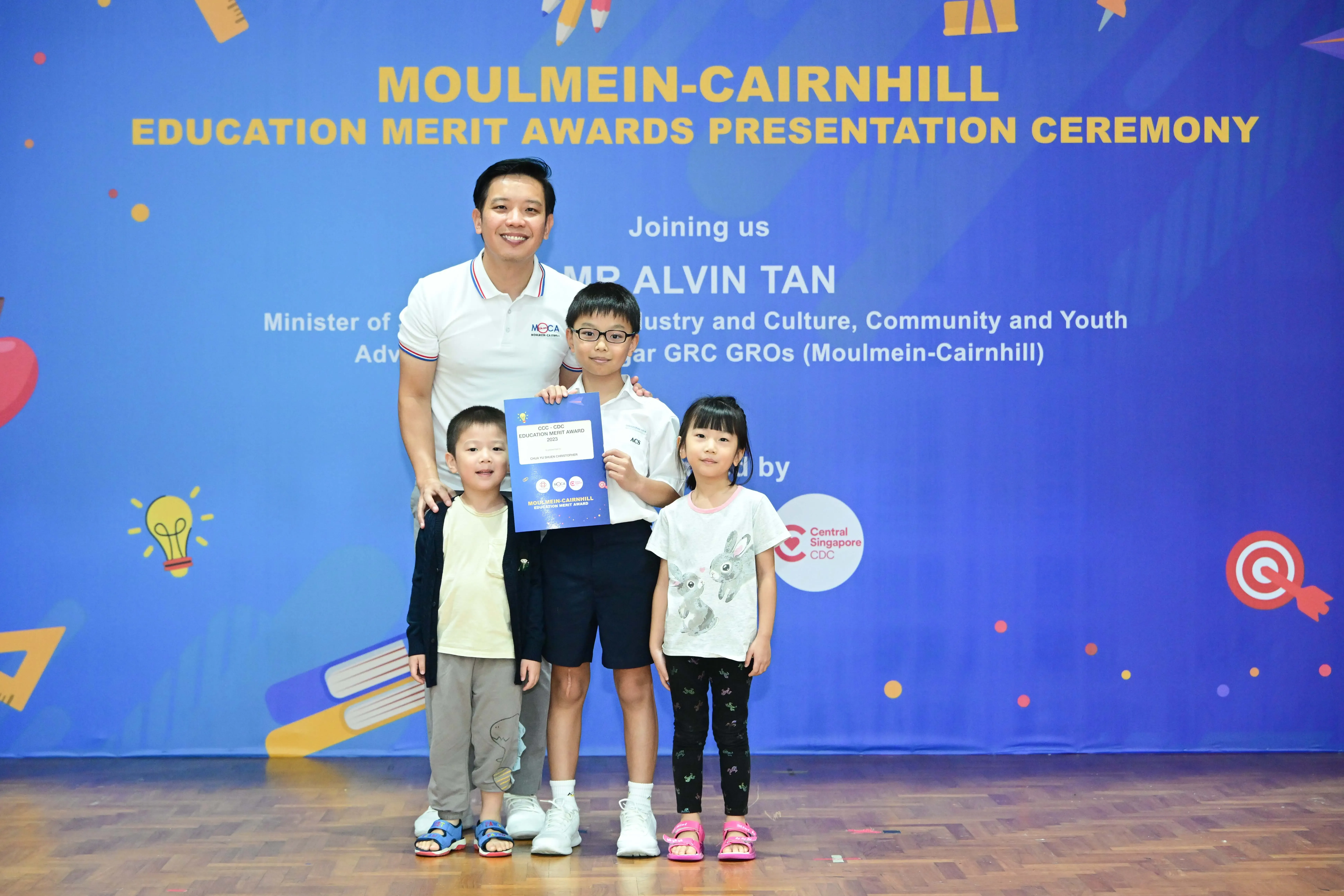 Man and three children pose before a Moulmein-Cairnhill Education Merit Awards Presentation Ceremony banner.