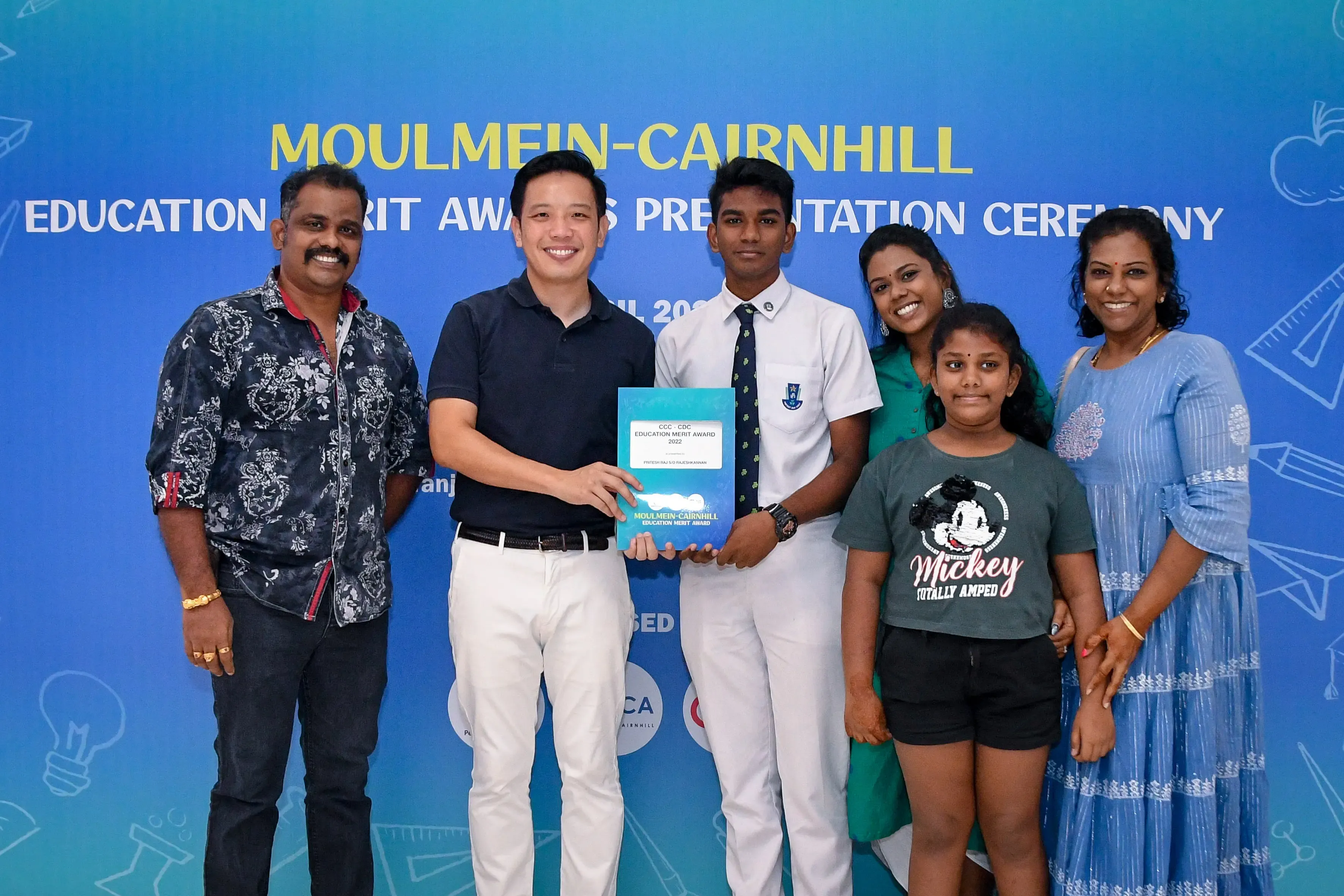 Six people pose smiling before a Moulmein-Cairnhill Education Merit Awards Presentation Ceremony banner.