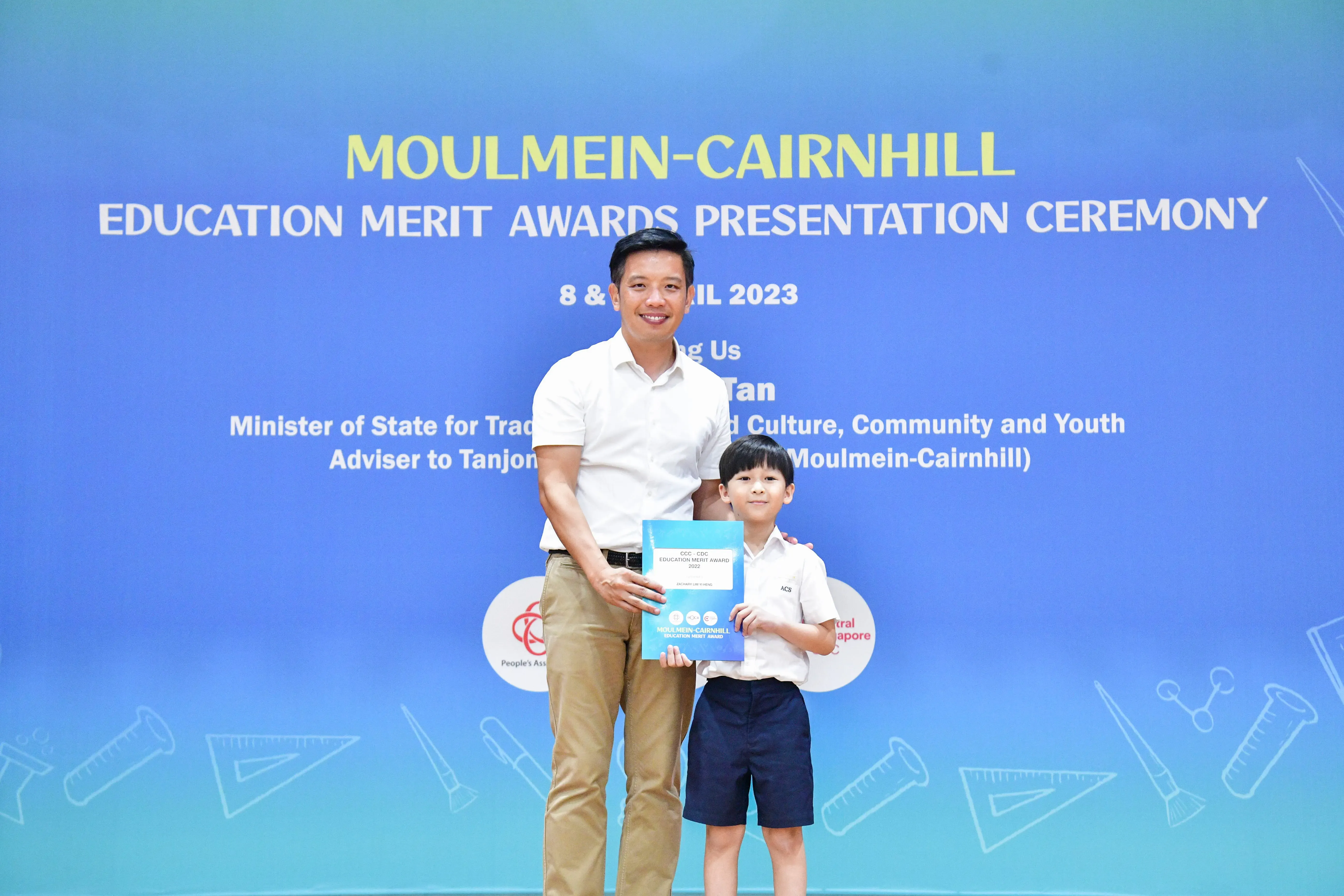 A man and a young boy hold a certificate in front of a Moulmein-Cairnhill Education Merit Awards 2023 banner.