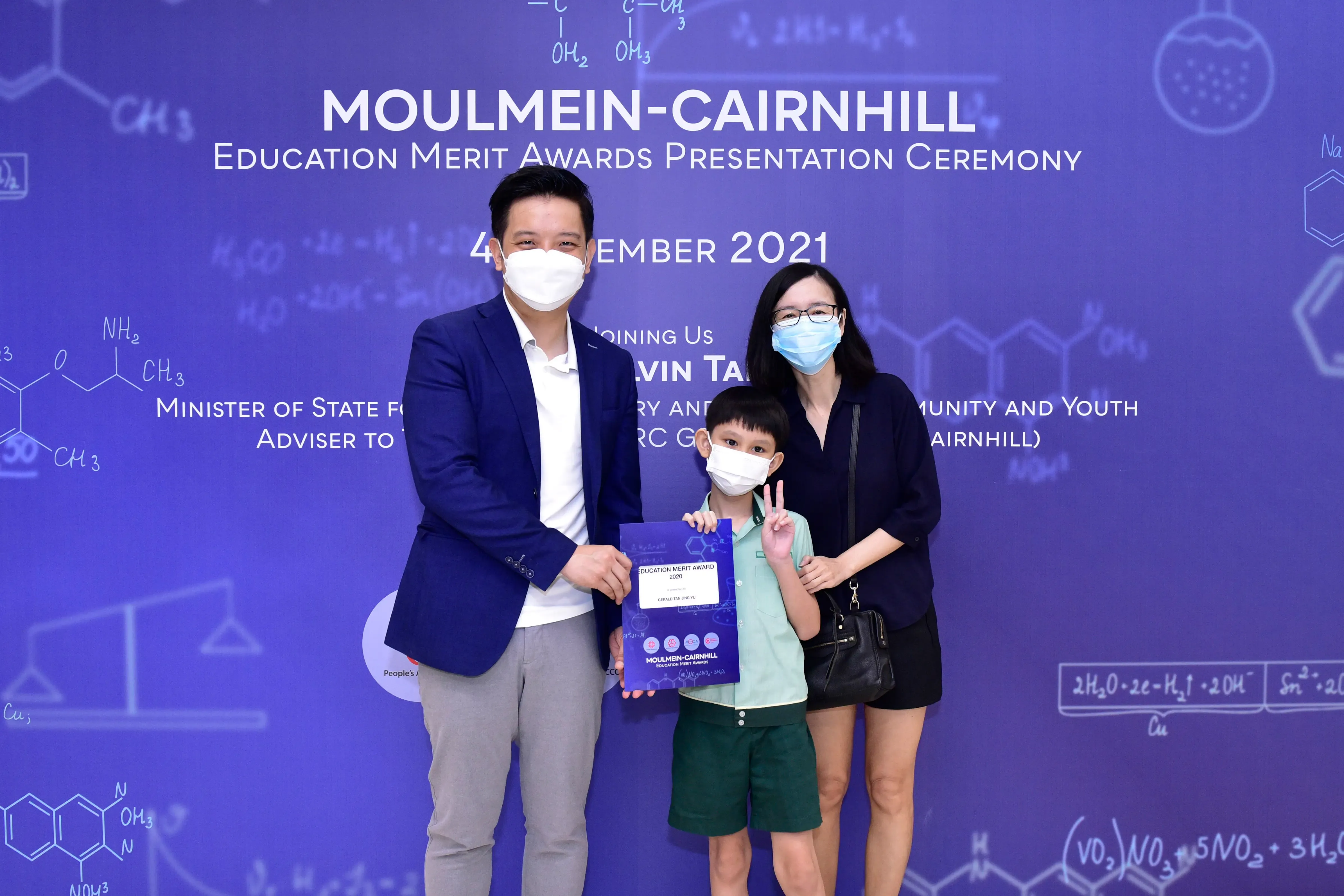 Two masked adults and a masked boy holding an award certificate pose at the Moulmein-Cairnhill Education Merit Awards 2021.