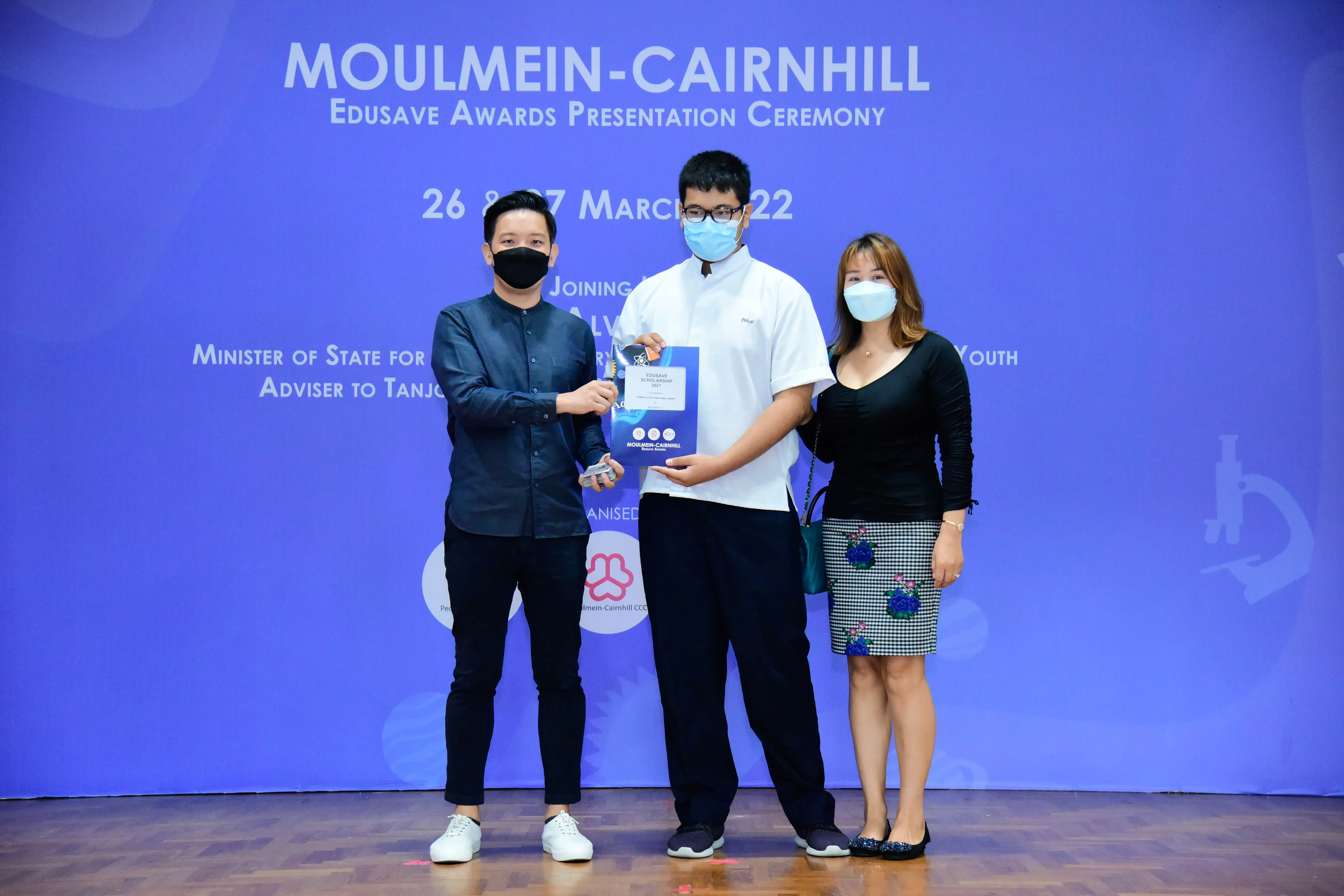 Three masked people stand on a stage at the Moulmein-Cairnhill Edusave Awards Presentation Ceremony, March 2022.