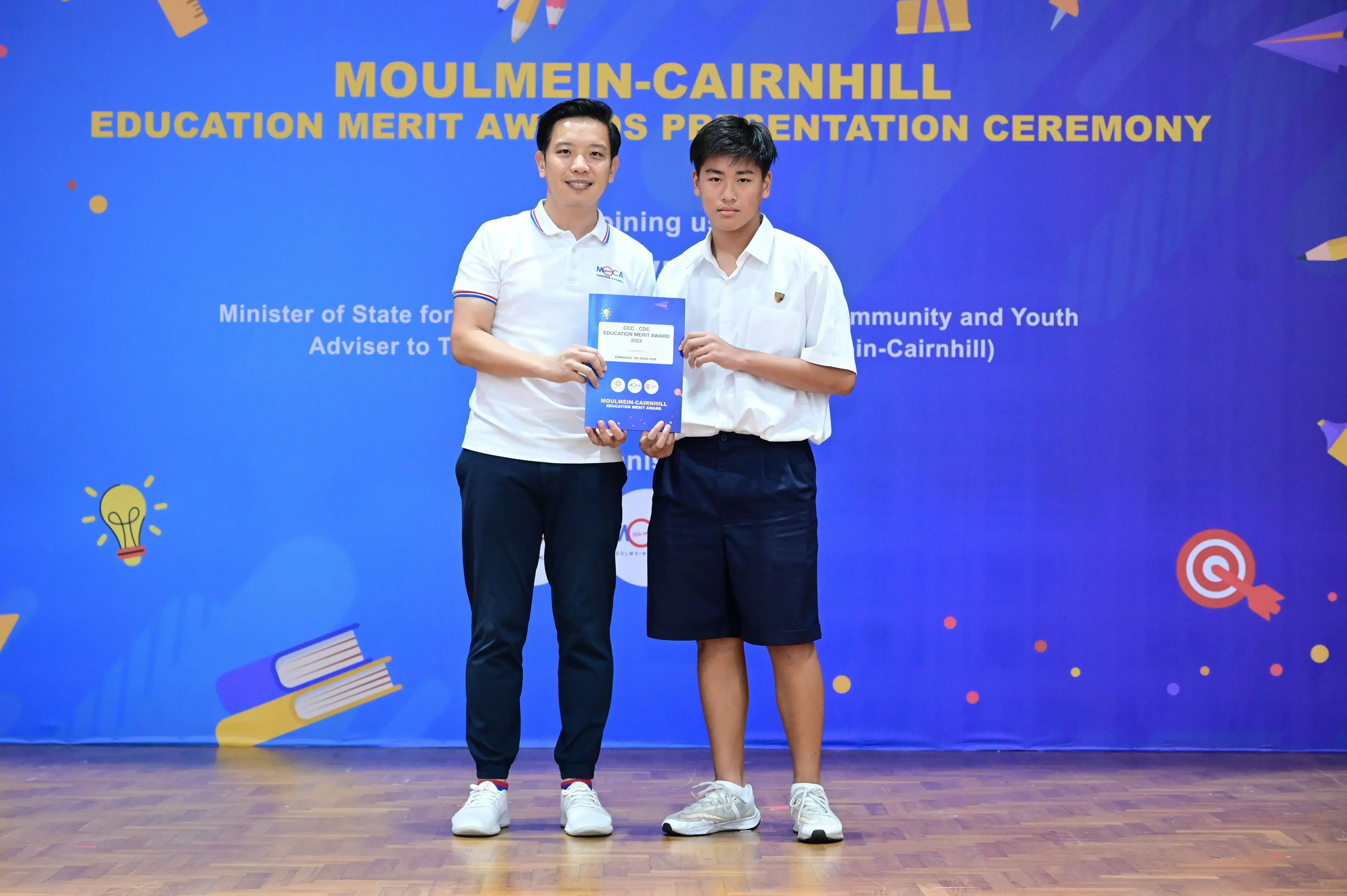 Two people hold a certificate together in front of a Moulmein-Cairnhill Education Merit Awards banner.