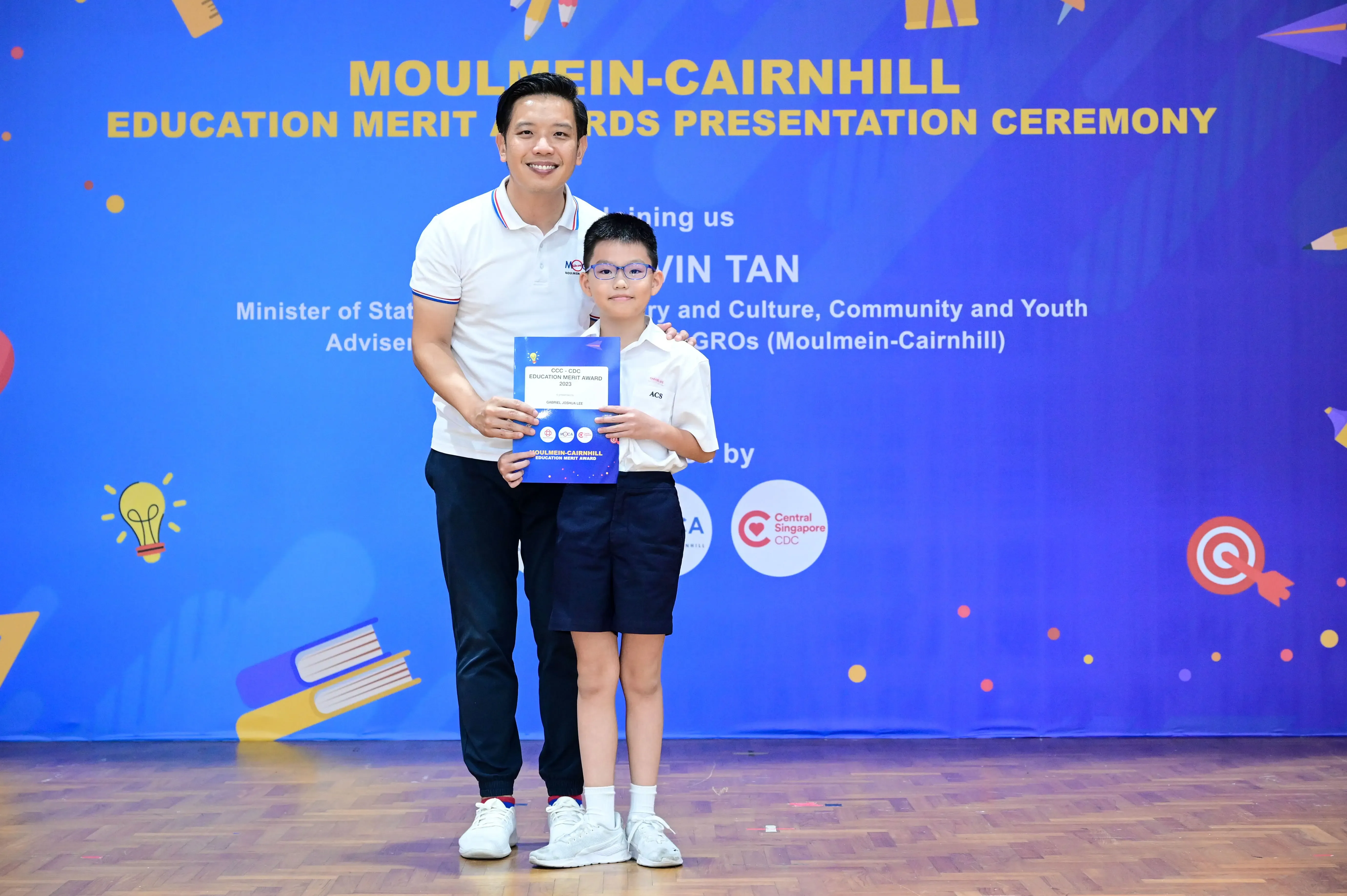 A man and a young boy holding an award certificate on stage at the Moulmein-Cairnhill Education Merit Awards Ceremony.