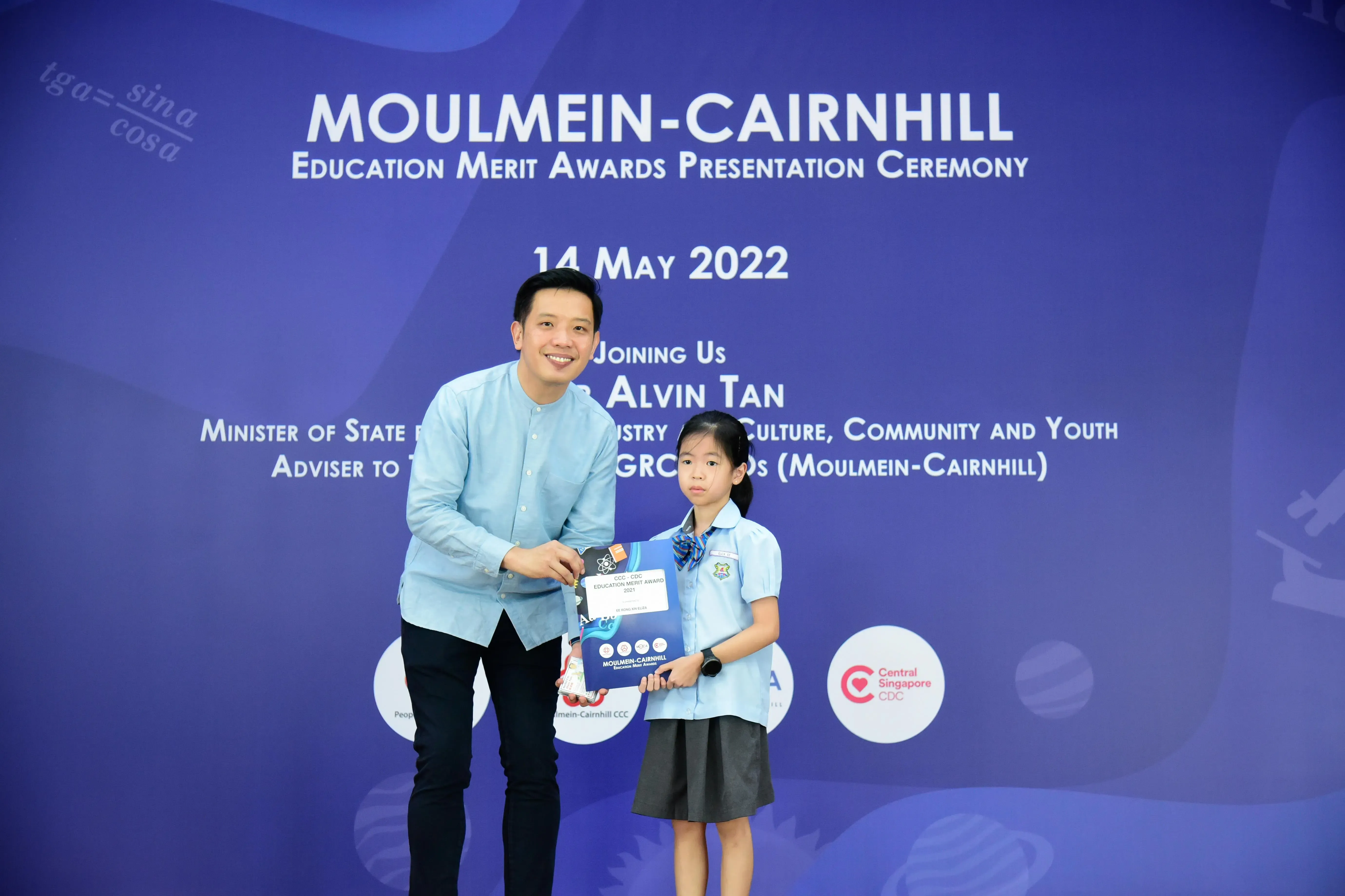 A man and a young girl in school uniform hold an award certificate at the Moulmein-Cairnhill Education Merit Awards ceremony.