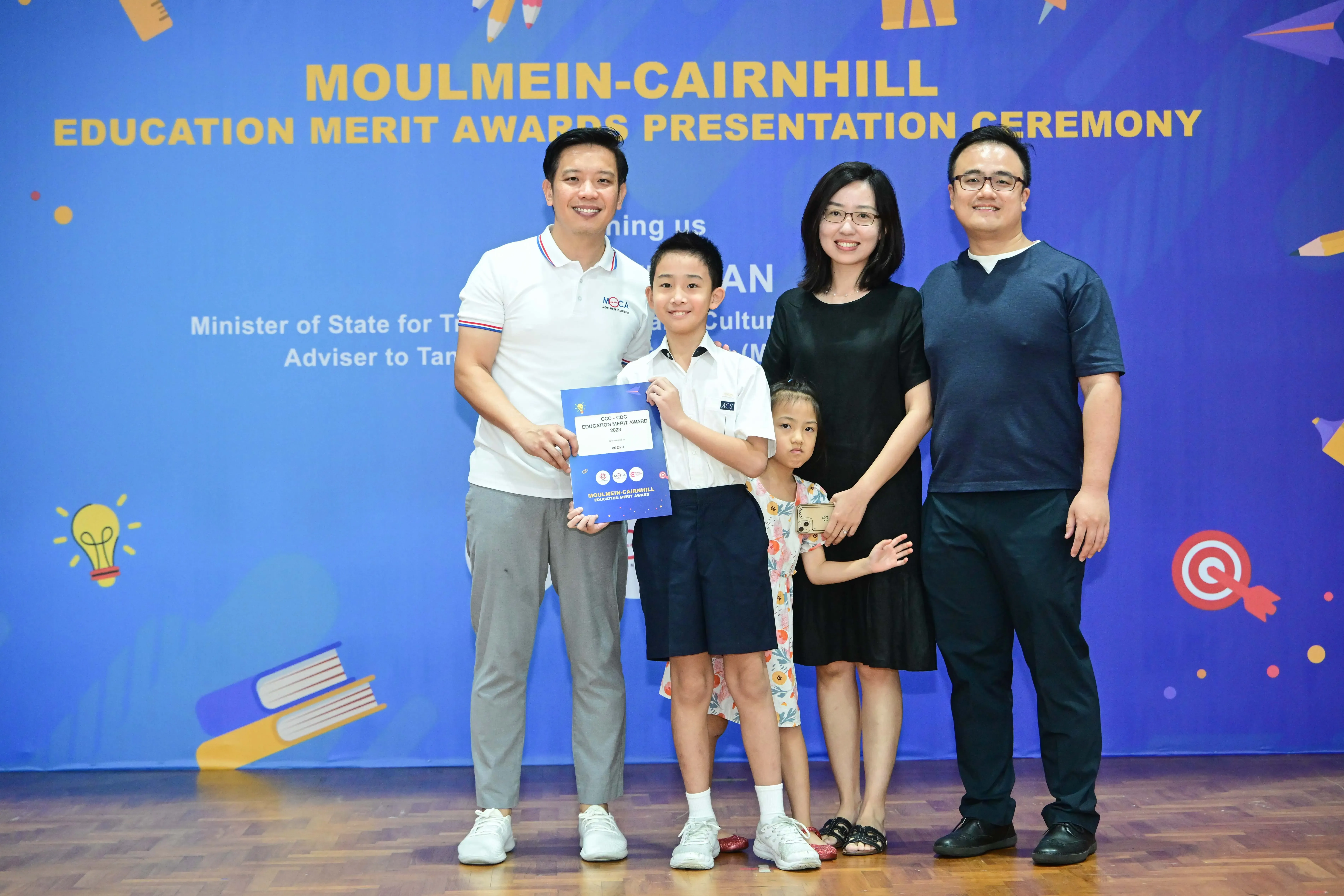 A boy holds an award certificate with three adults and a young girl at the Moulmein-Cairnhill Education Merit Awards ceremony.