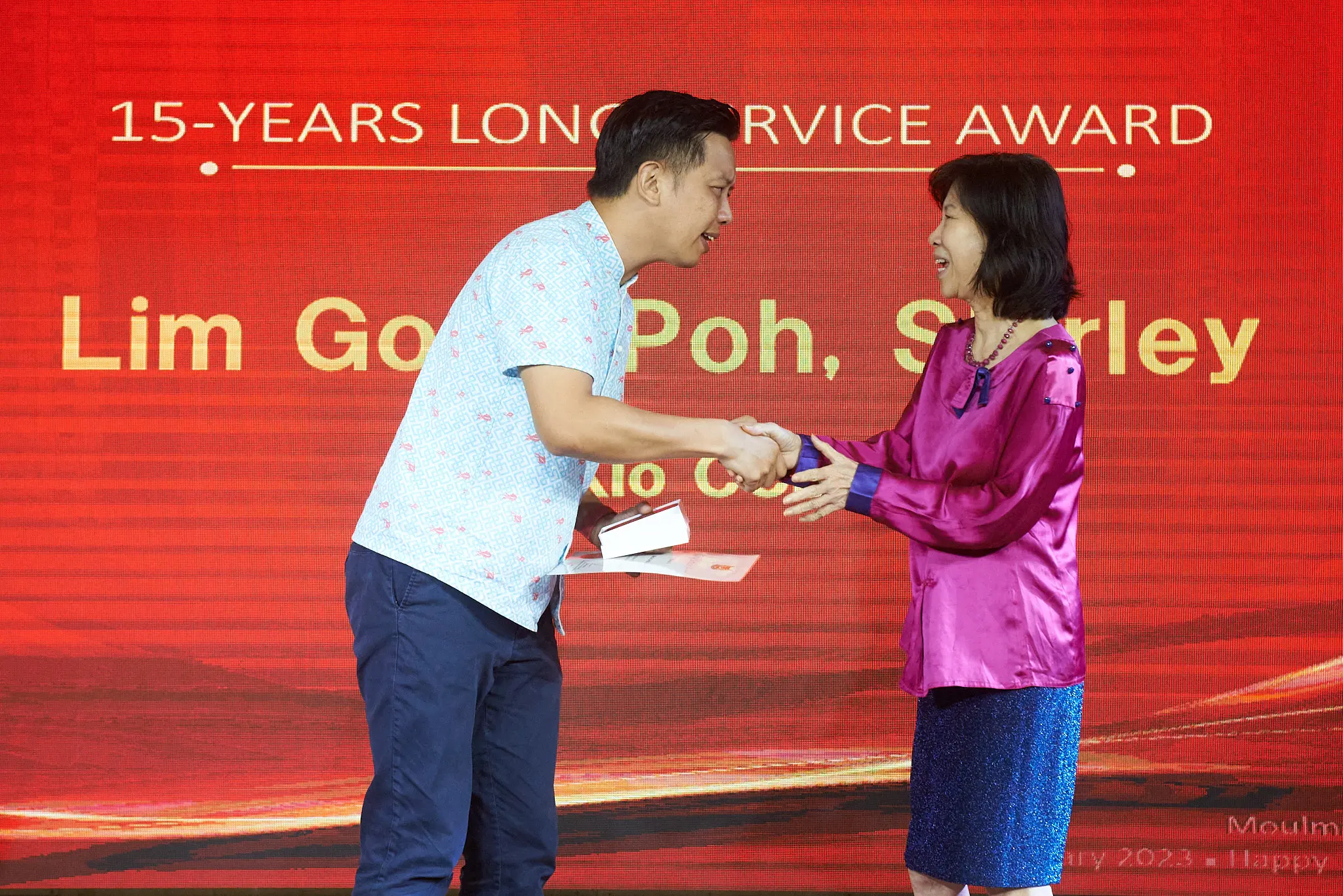 Two people shake hands on stage in front of a red screen displaying a 15-Years Long Service Award.