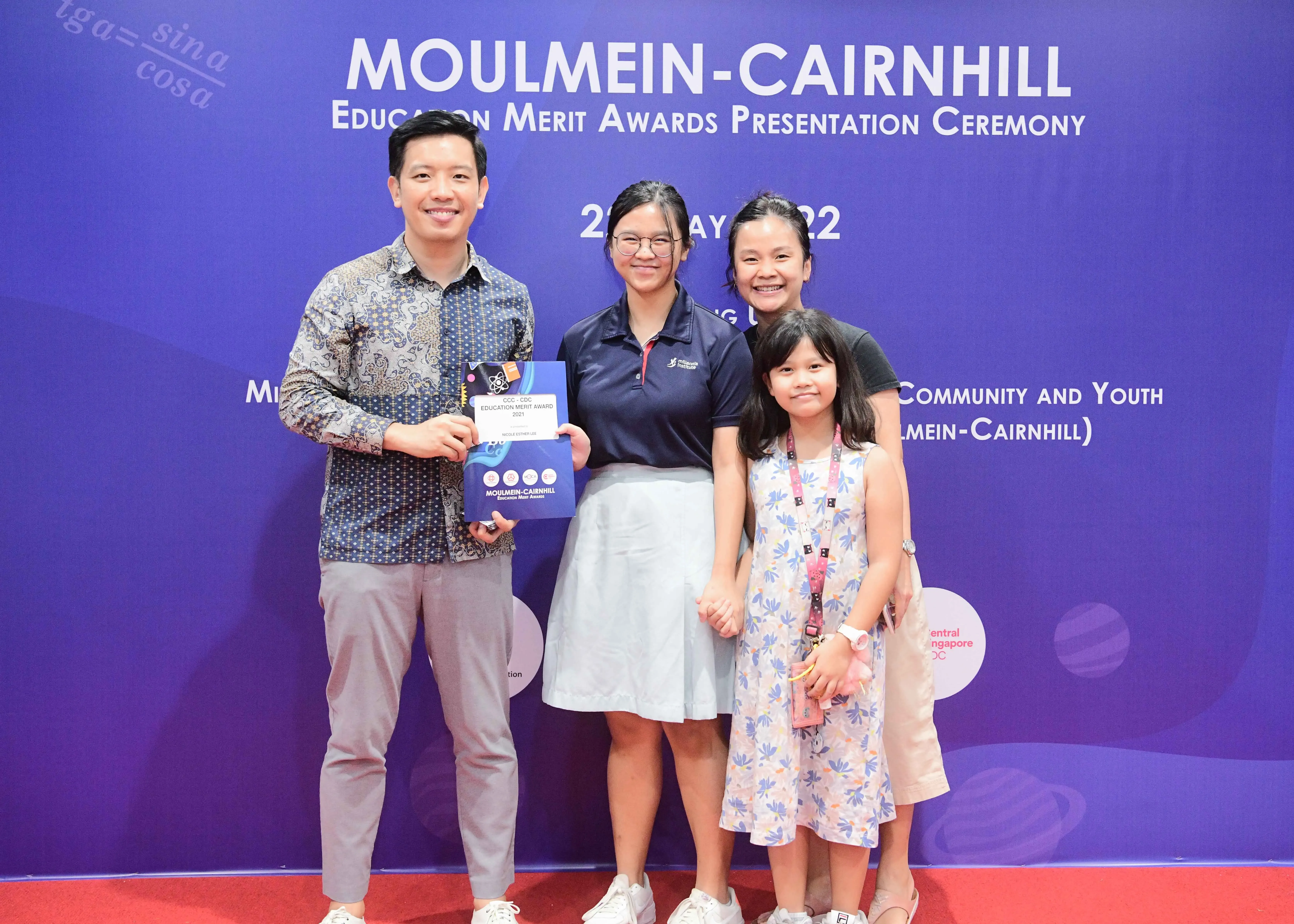 Four people pose smiling before a Moulmein-Cairnhill Education Merit Awards Presentation Ceremony banner.