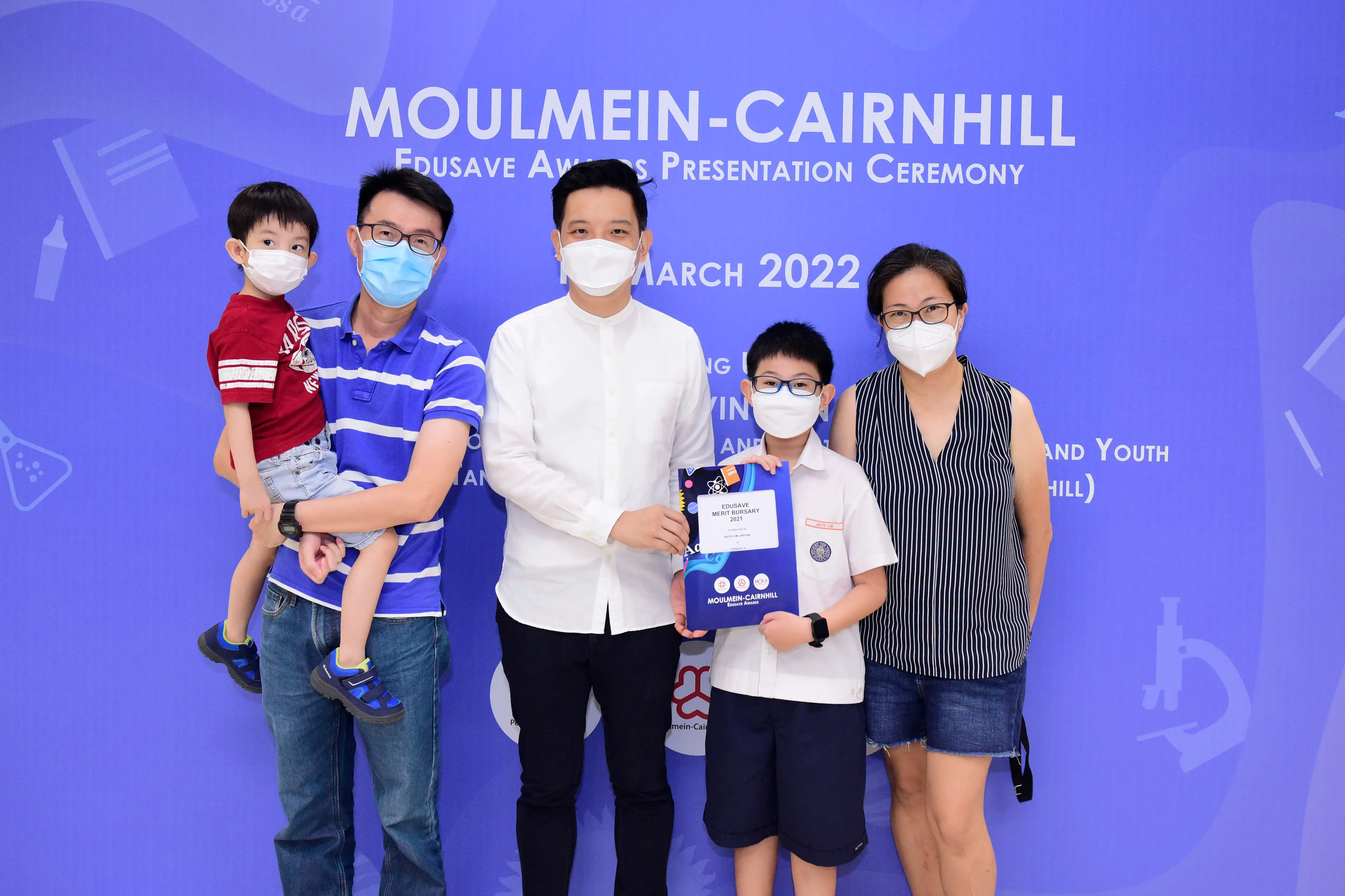 Five masked people pose at the Moulmein-Cairnhill Edusave Awards Presentation Ceremony, March 2022, holding a bursary certificate.