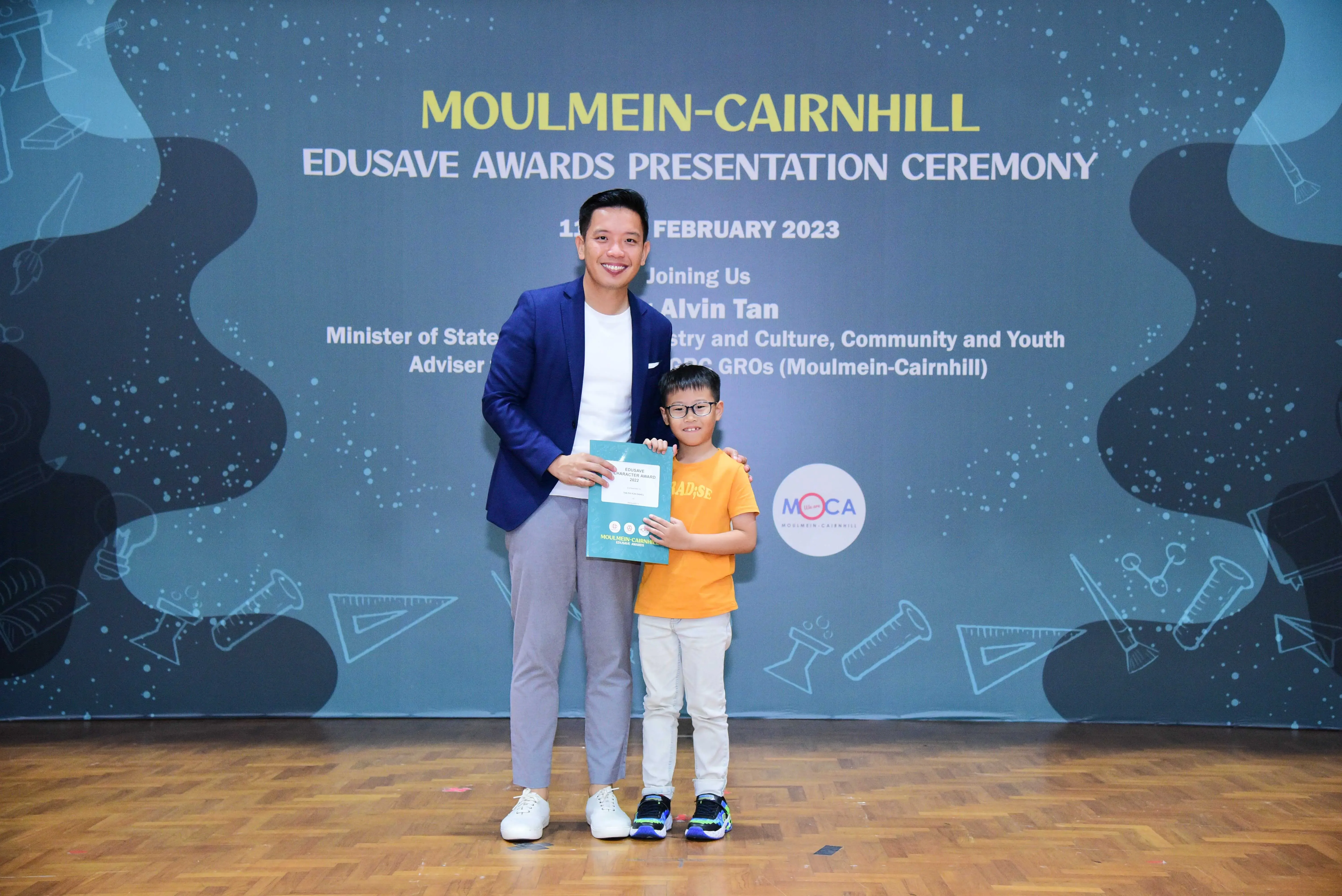 An adult in a blue blazer and a young boy in a yellow shirt hold a certificate at the Moulmein-Cairnhill Edusave Awards 2023.