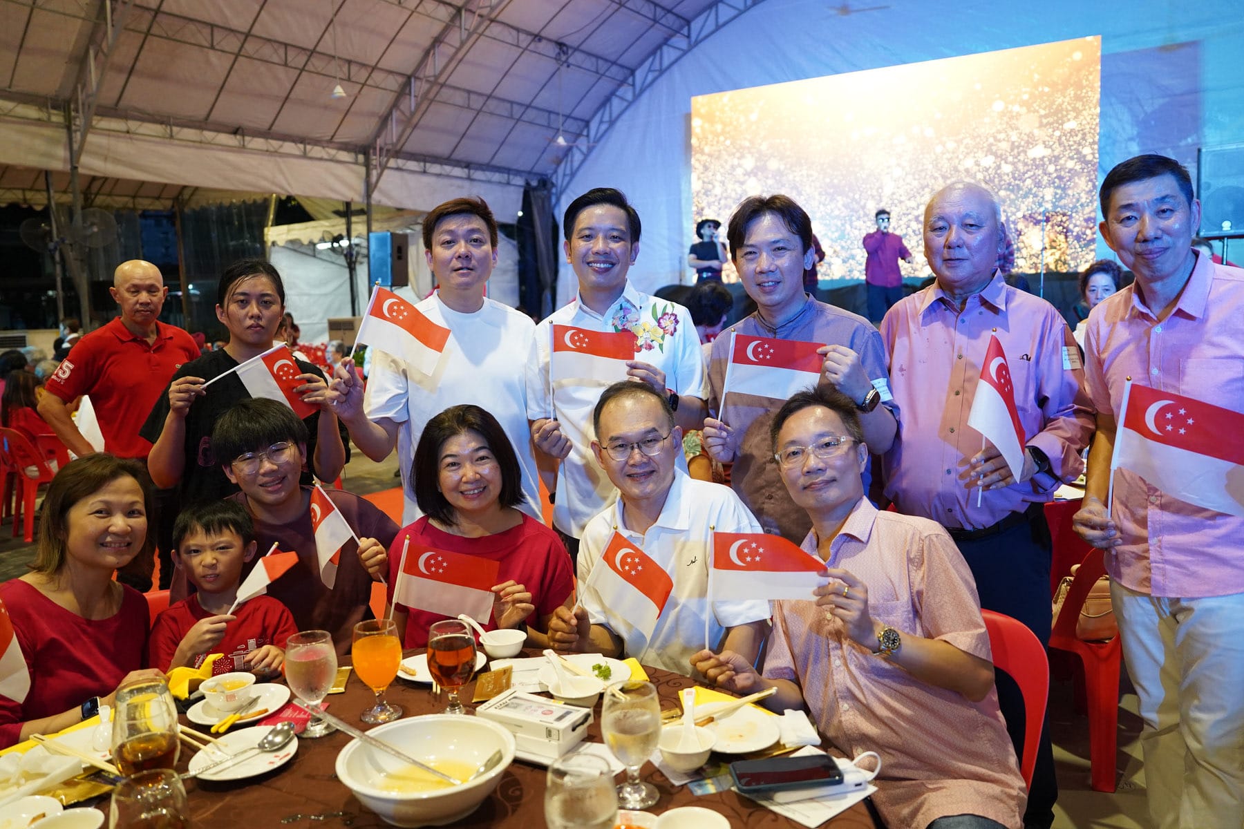 A group of smiling people holding small Singapore flags at an outdoor dining event under a tent.