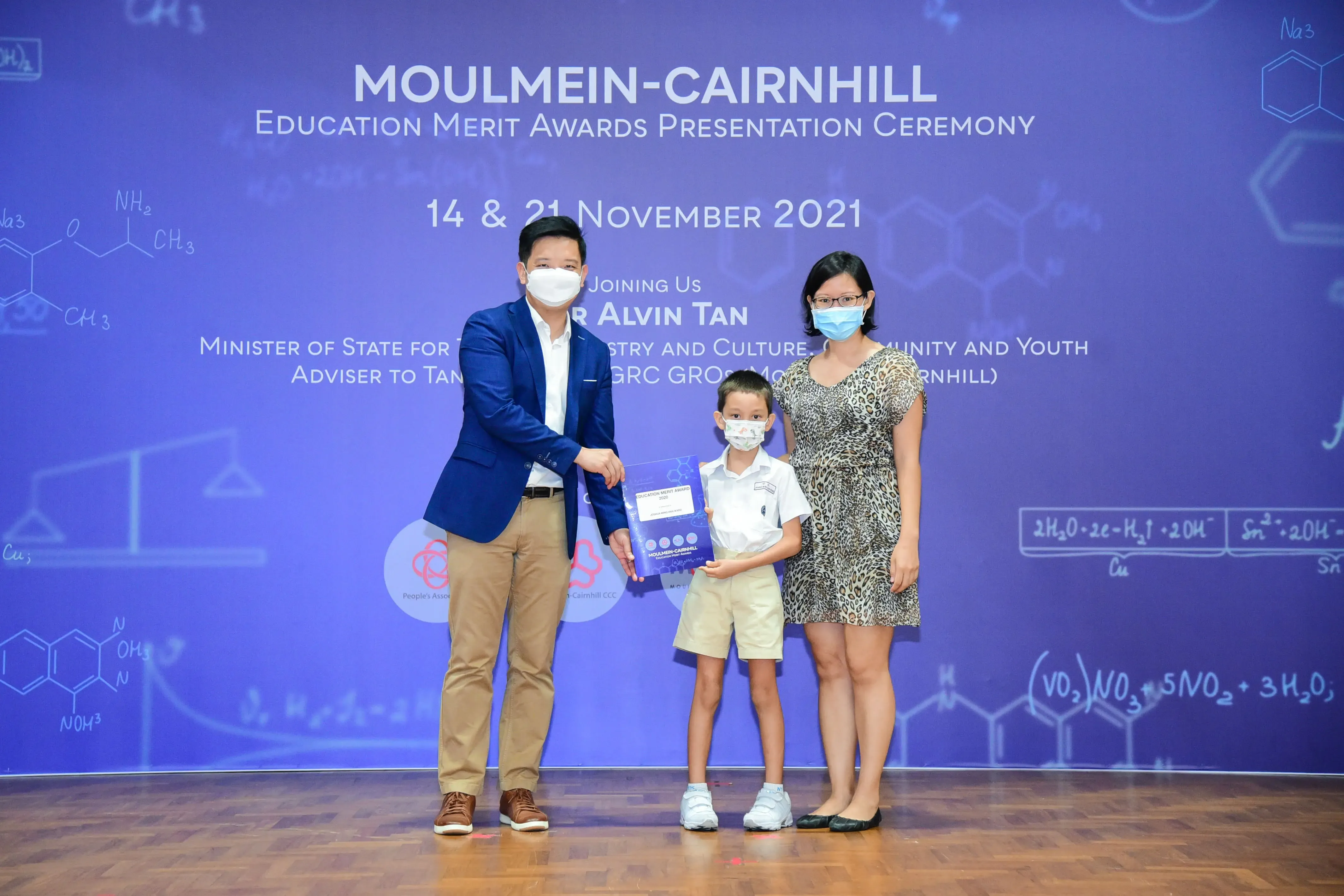 A man presents an award to a young boy with a woman beside them at the Moulmein-Cairnhill Education Merit Awards 2021.