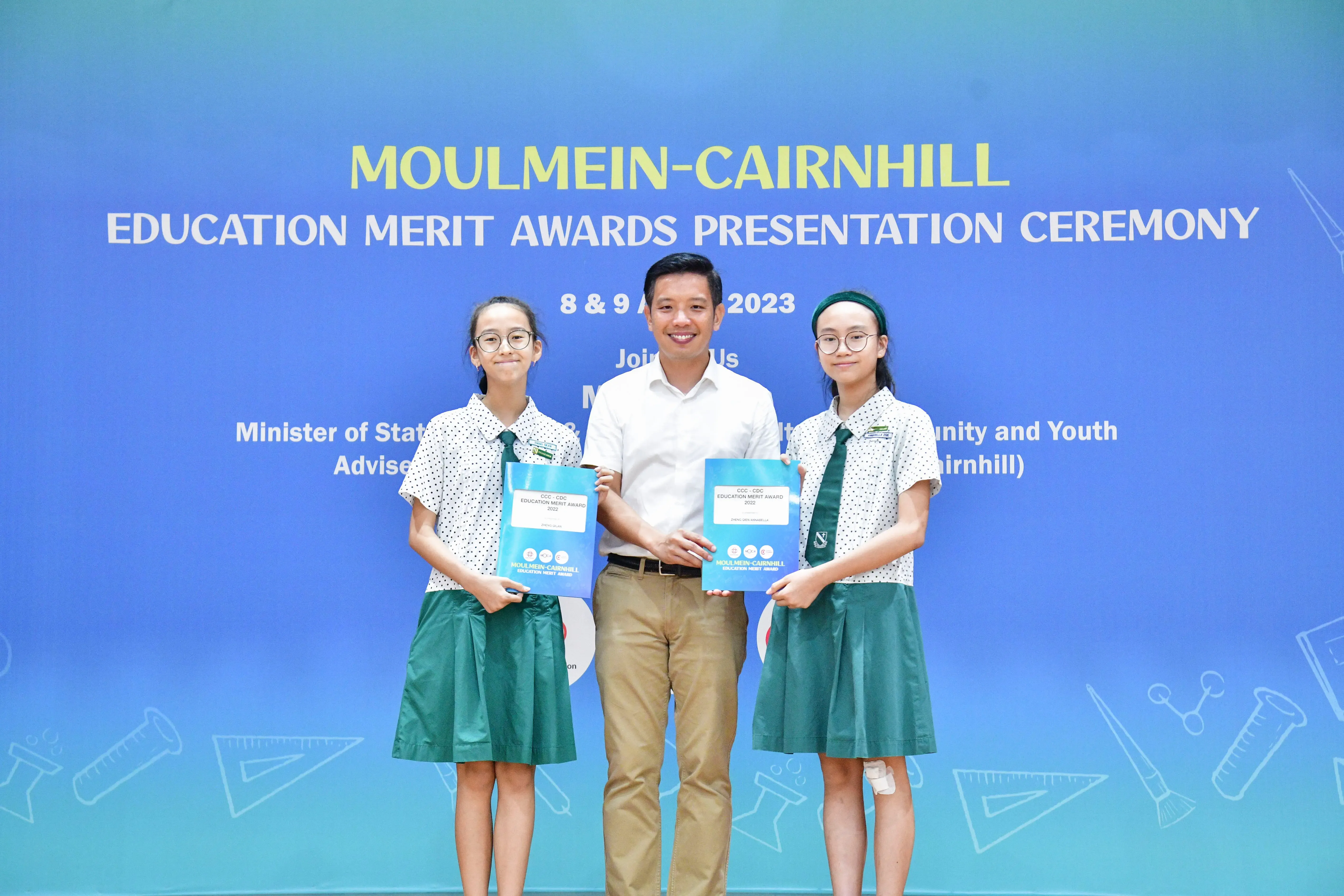 A man stands between two uniformed schoolgirls holding award folders at the Moulmein-Cairnhill Education Merit Awards 2023.