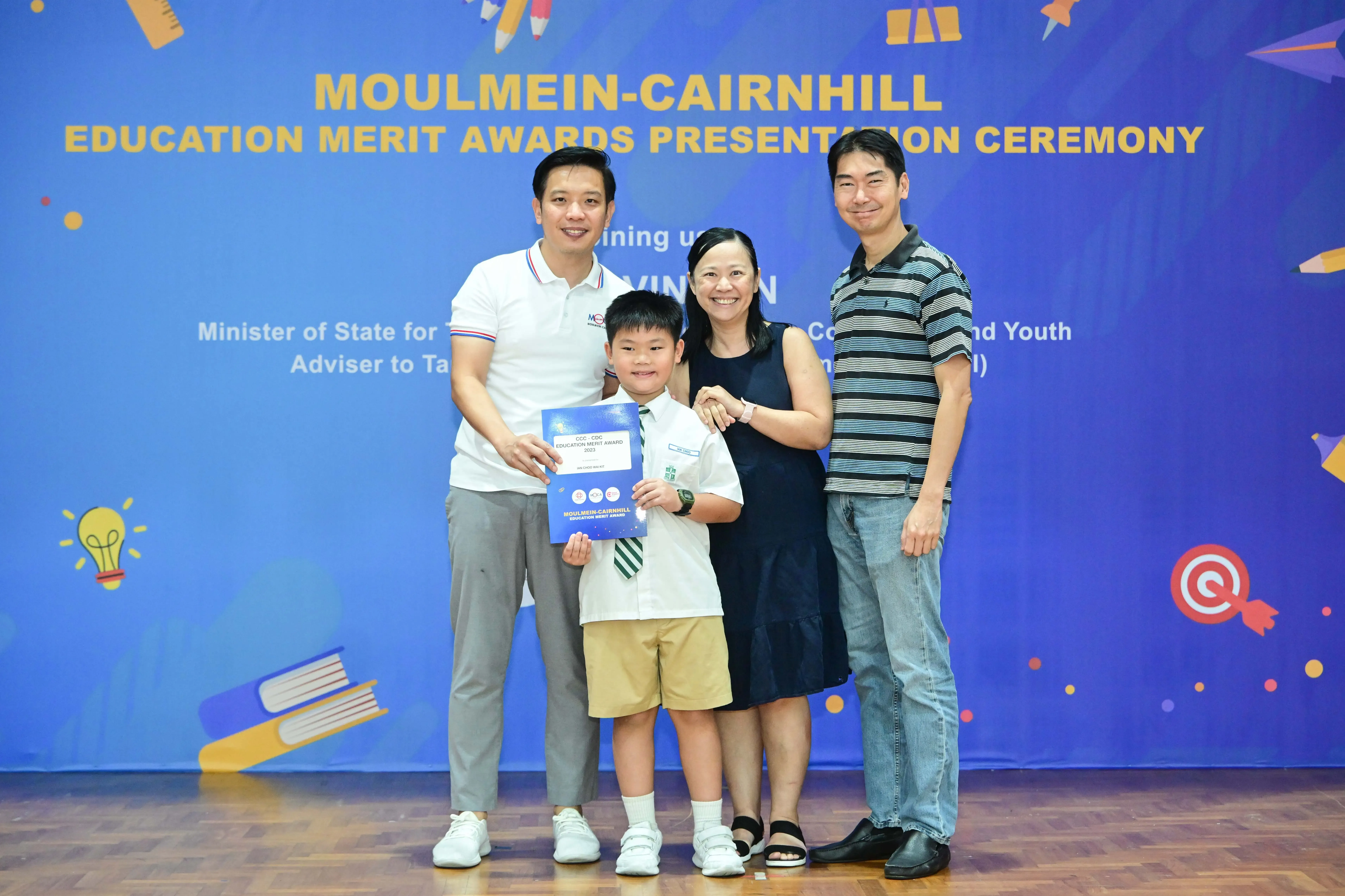 Three adults and a young boy in school uniform hold a certificate at the Moulmein-Cairnhill Education Merit Awards ceremony.