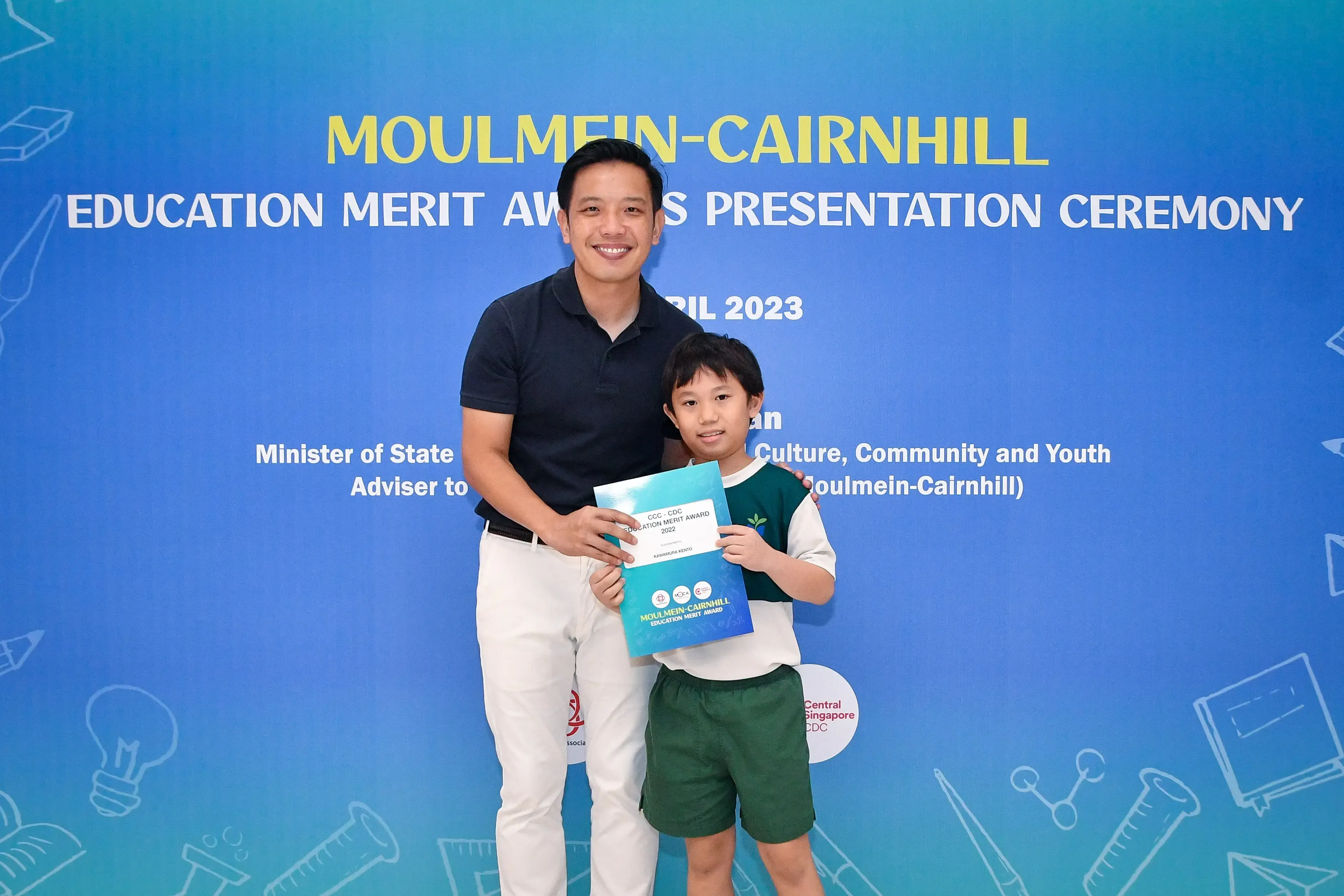 Man and young boy hold an award certificate at the Moulmein-Cairnhill Education Merit Awards Presentation Ceremony 2023.