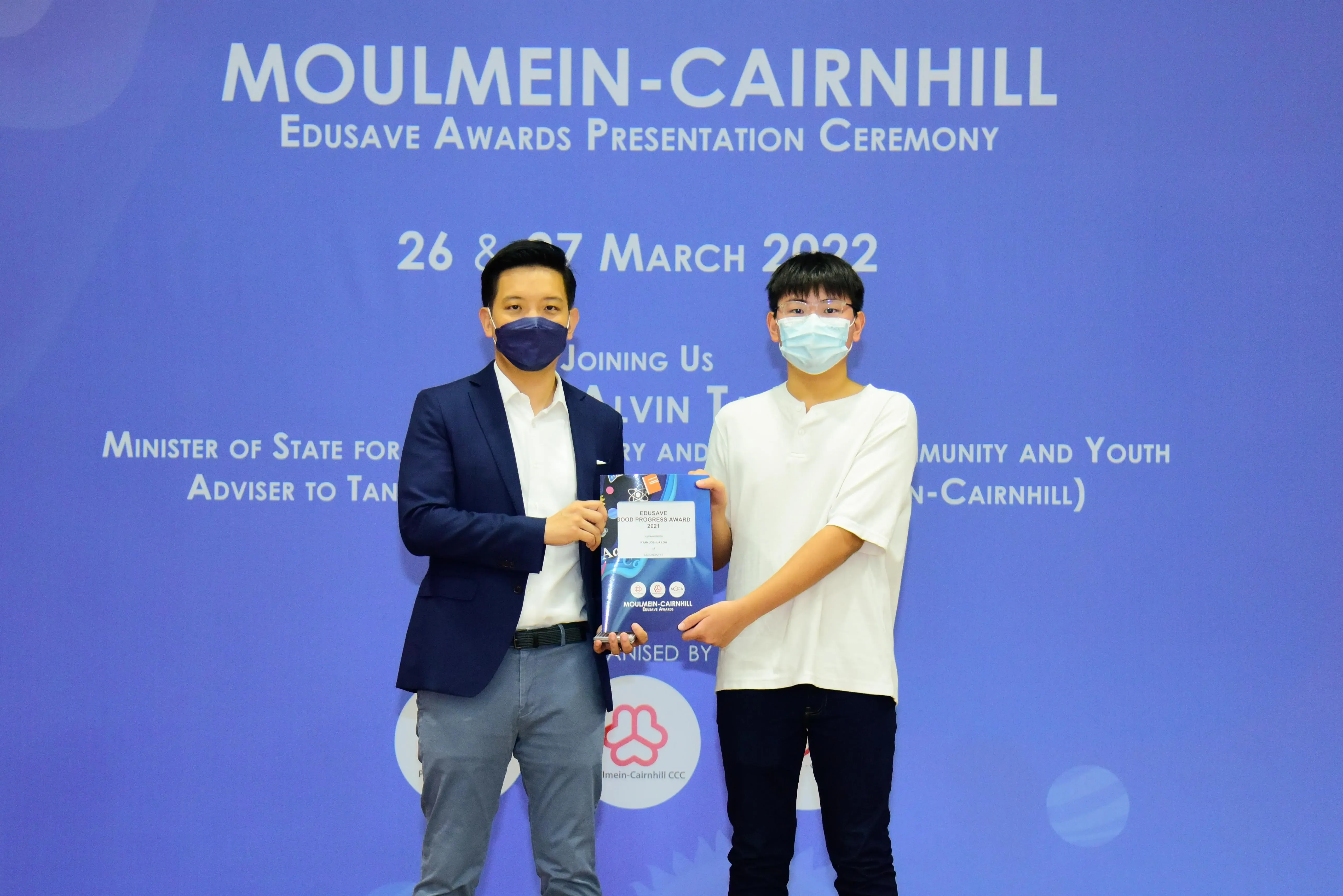 Two masked men hold an Edusave award at the Moulmein-Cairnhill Edusave Awards Presentation Ceremony, March 2022.