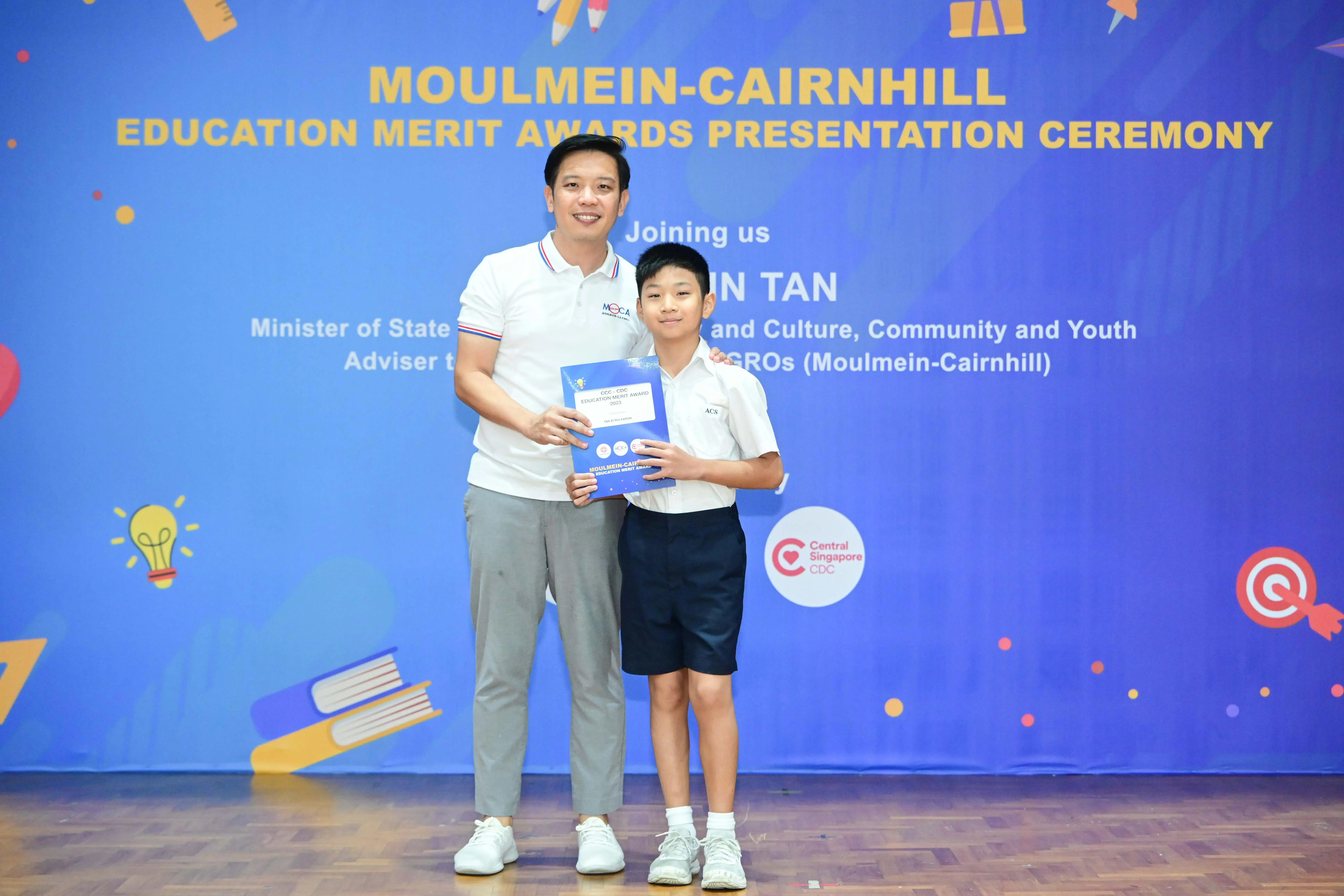 Adult and young boy holding an award booklet at Moulmein-Cairnhill Education Merit Awards Presentation Ceremony.