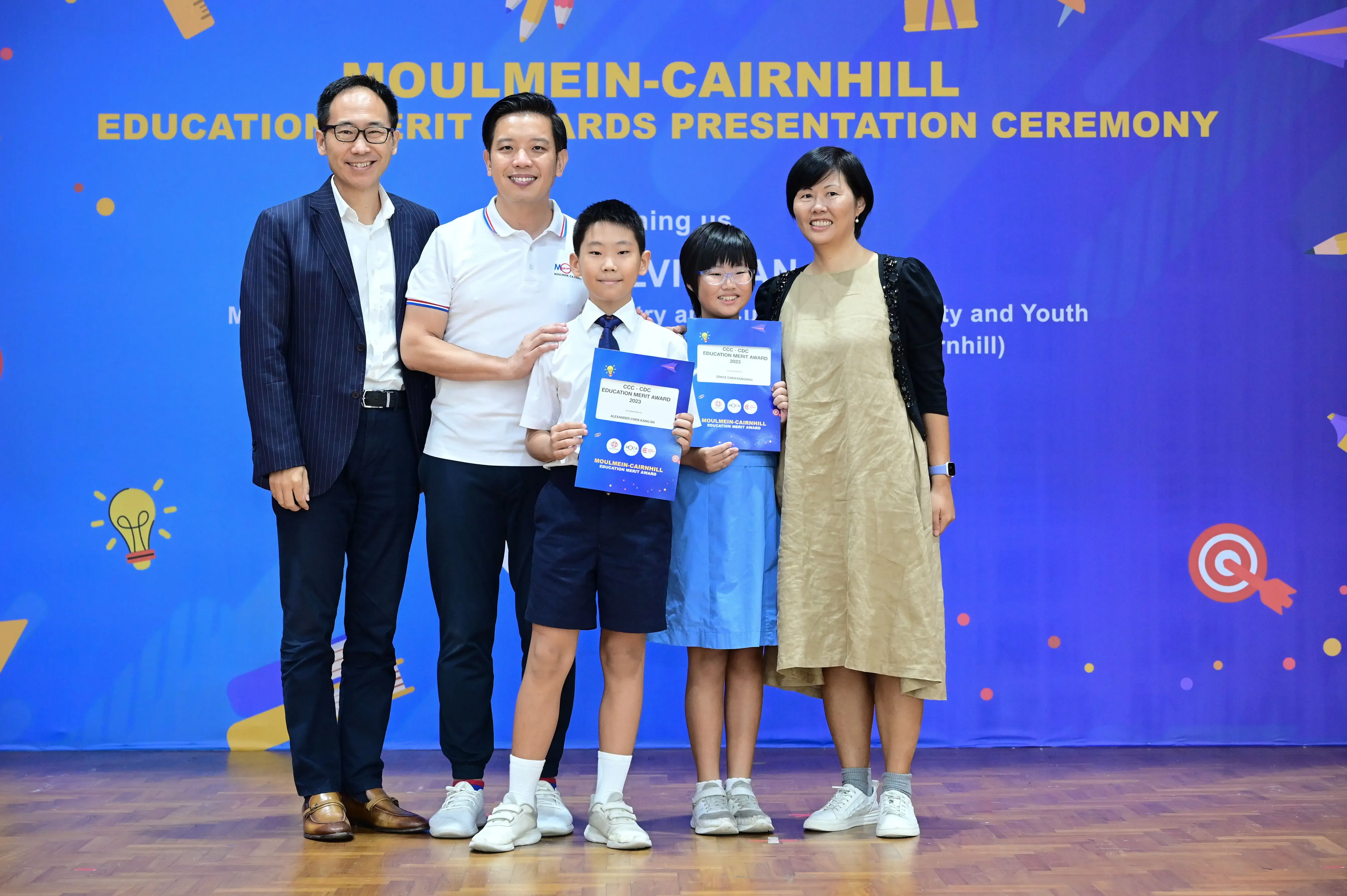 Five people pose at the Moulmein-Cairnhill Education Merit Awards ceremony, two children holding award certificates.
